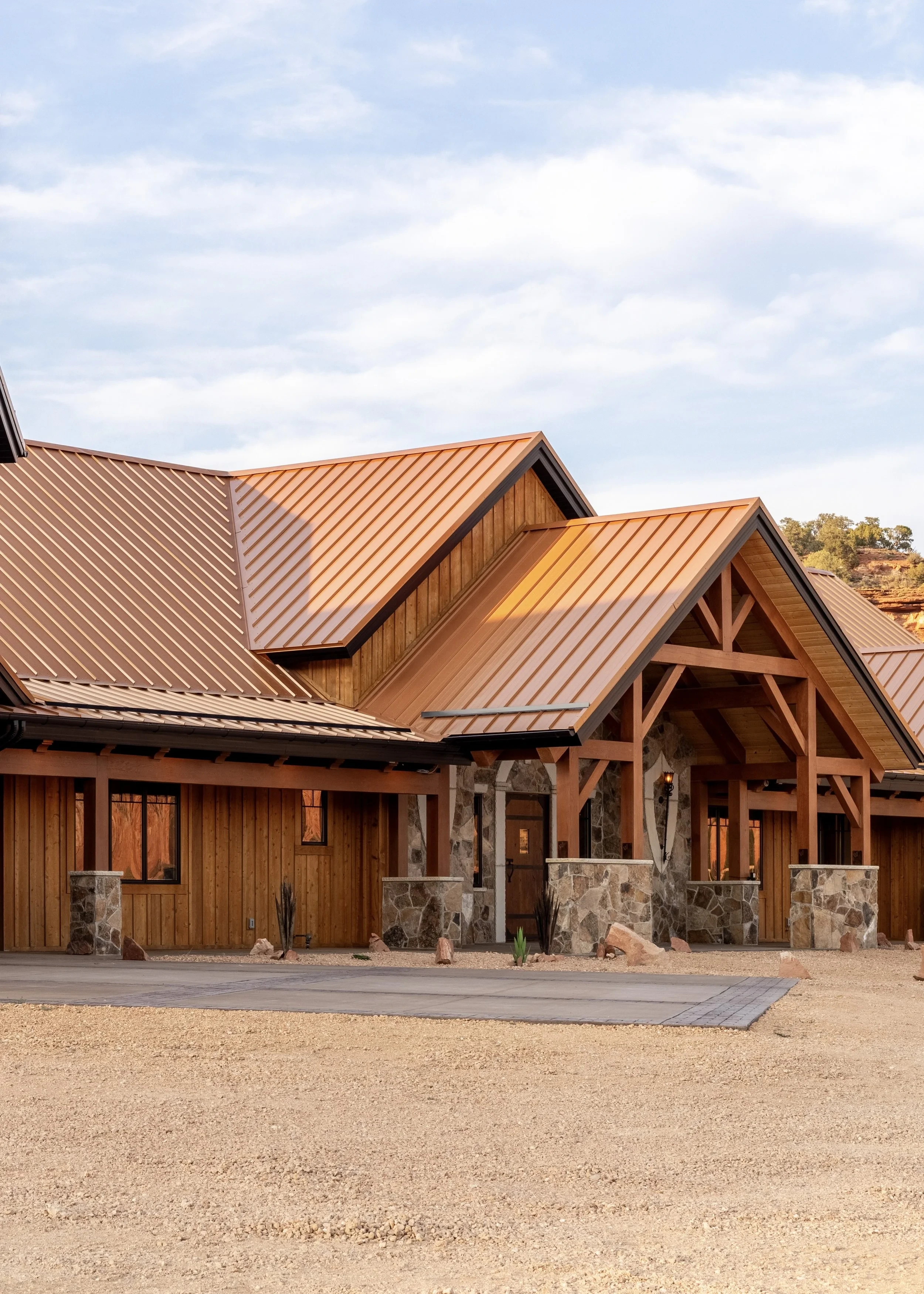 A modern house with wooden and stone exterior, with a copper roof, situated on a gravel lot under a partly cloudy sky.