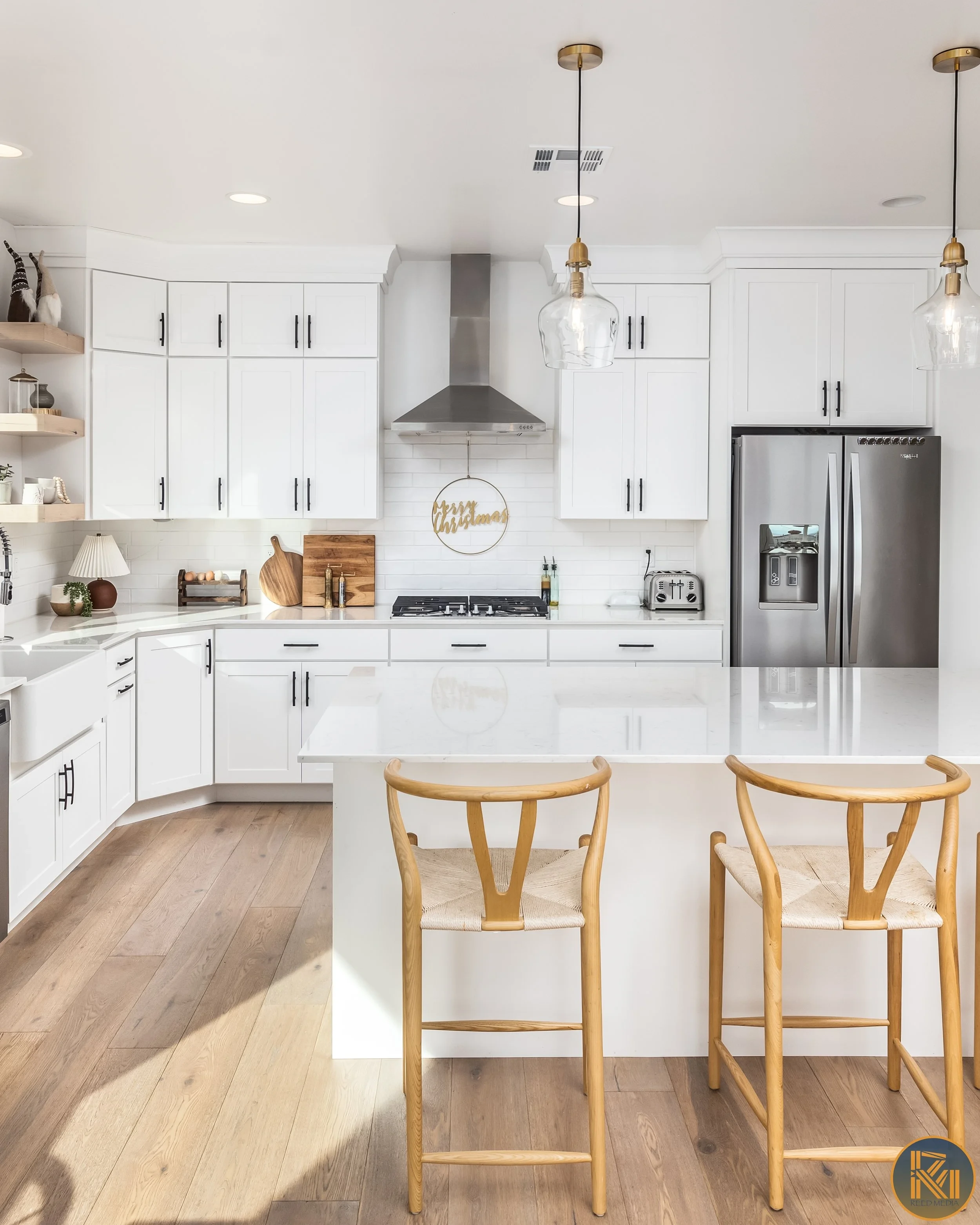 Modern white kitchen with island, two wooden chairs, stainless steel refrigerator, range and vent, white cabinets, and shelves with decorations, holiday sign, and kitchen utensils.