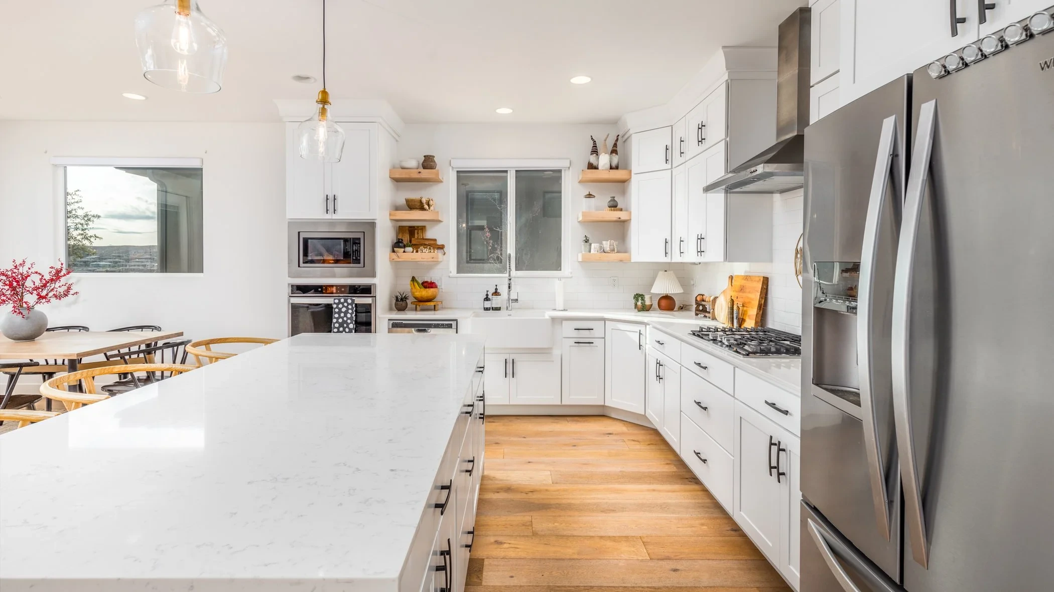 Modern kitchen with white cabinets, large island with white countertop, stainless steel refrigerator, stove, microwave, open shelving with decor, hardwood flooring, windows with natural light.