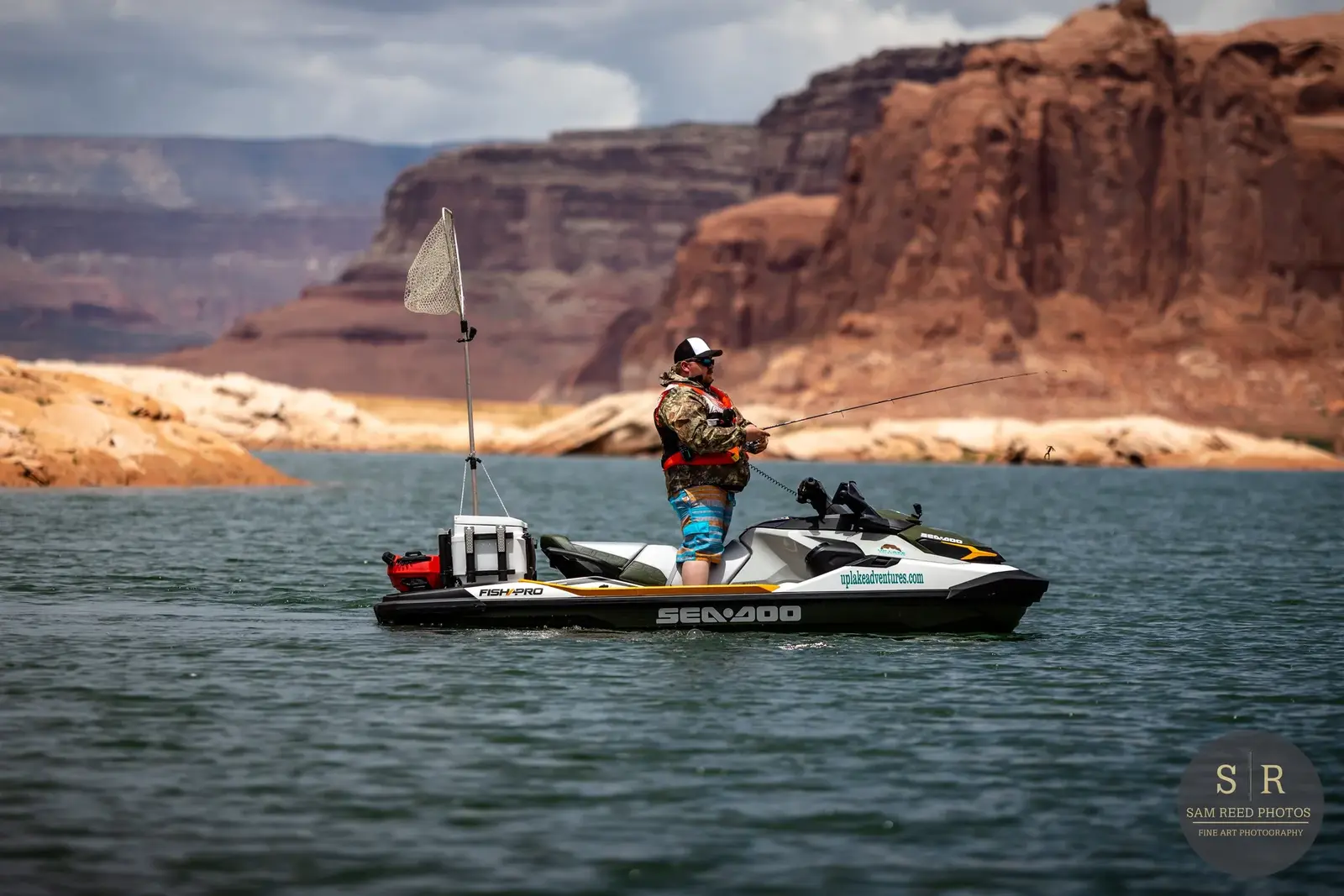 A person fishing on a Sea-Doo jet ski on a lake surrounded by red rock formations. The person is wearing a camouflage jacket, shorts, and a white cap.