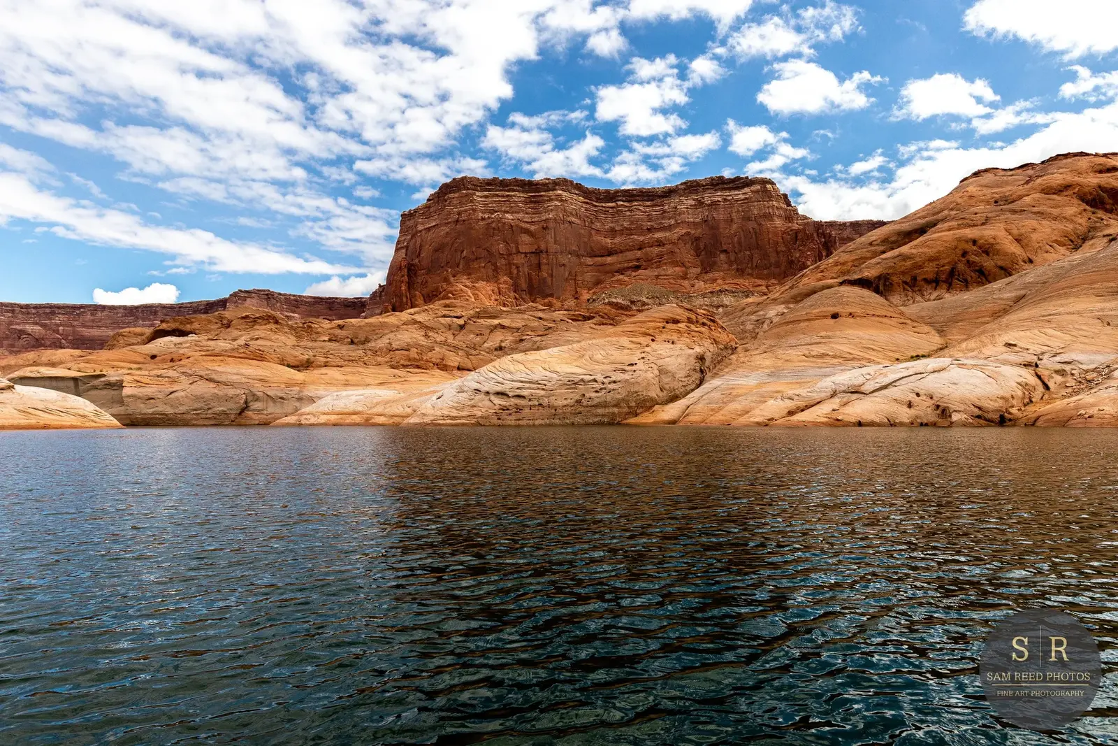 A serene body of water with ripples, surrounded by arid, layered red and tan rock formations under a blue sky with scattered white clouds.