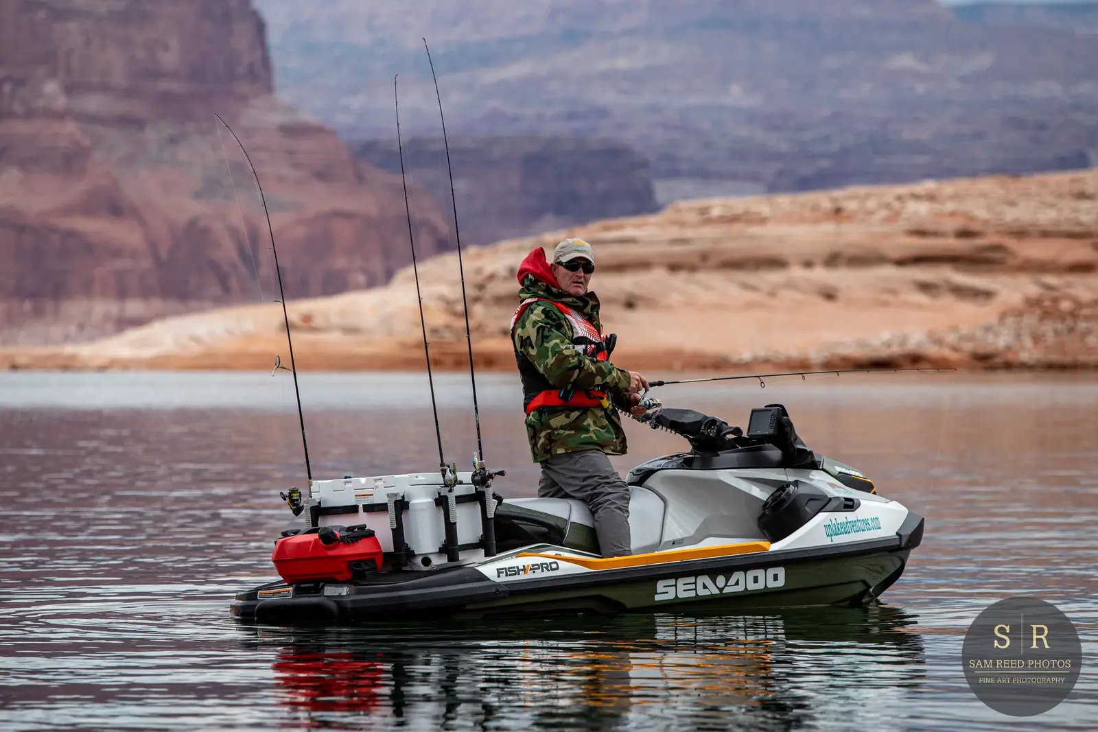 Man in camouflage jacket and hat fishing from a small boat on a calm lake with canyon cliffs in the background.