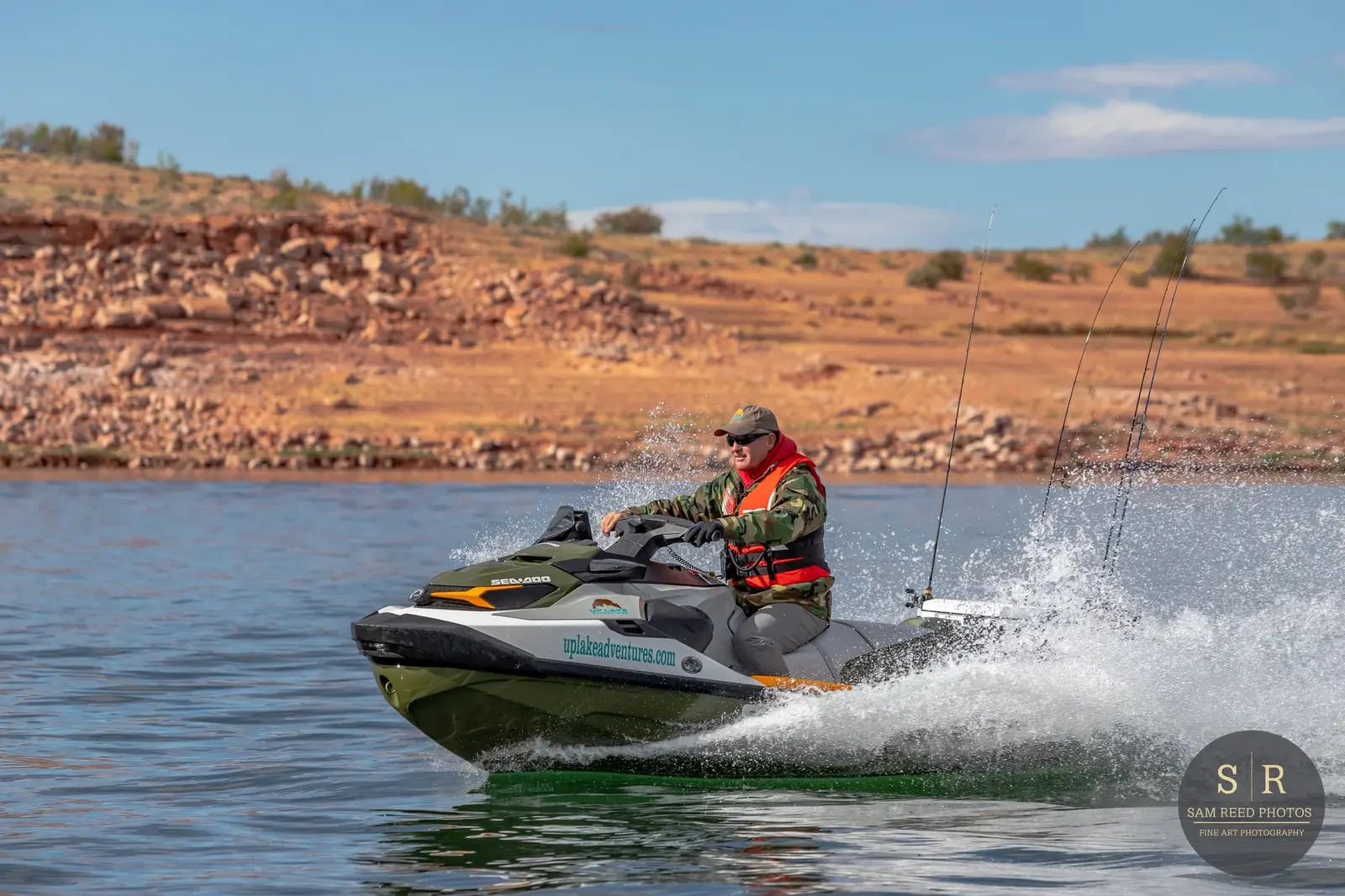 Man riding a black and green personal watercraft on a lake with a rocky, reddish-brown shoreline and a partly cloudy sky in the background.