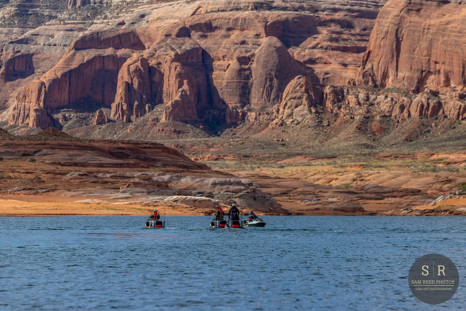 Four people fishing on boats in a lake with a backdrop of red rock formations and mountains in a desert landscape.