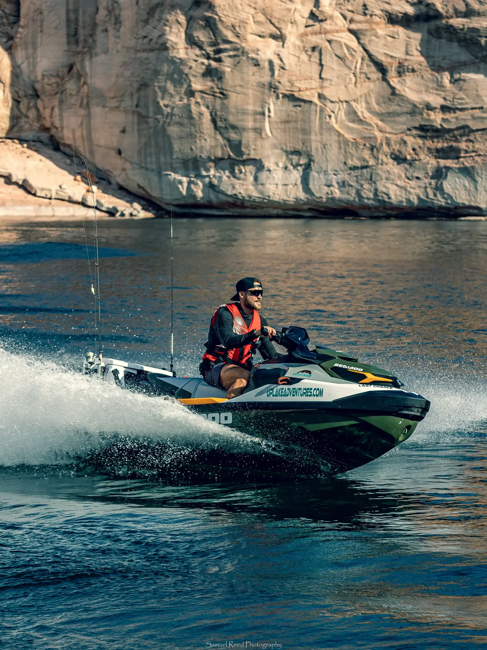 A man wearing sunglasses, a black cap, a red life jacket, and black shorts riding a Sea-Doo jet ski on a body of water with rocky cliffs in the background.