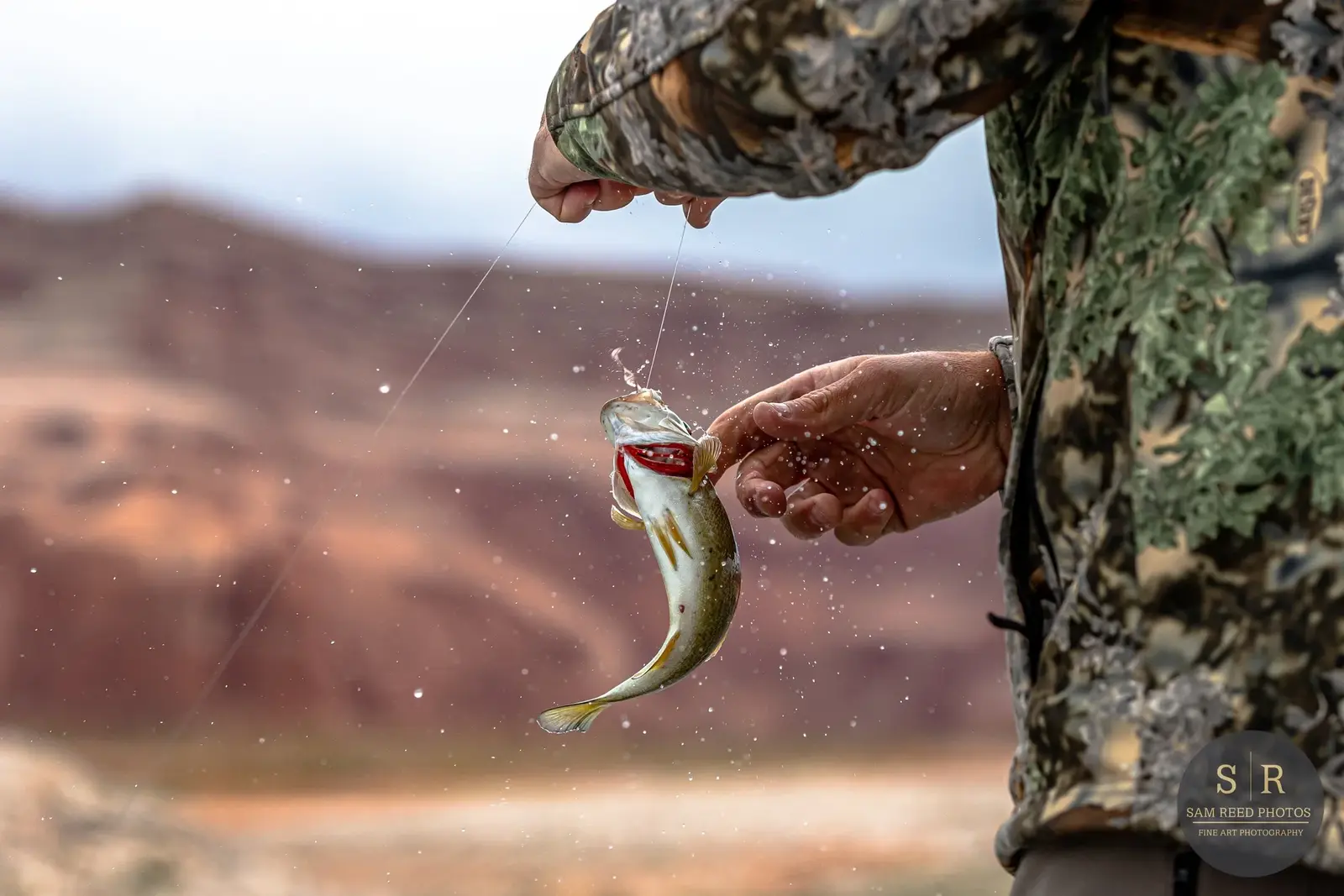 A person in camouflage clothing is fishing and holding a small fish above water, with water droplets splashing around.