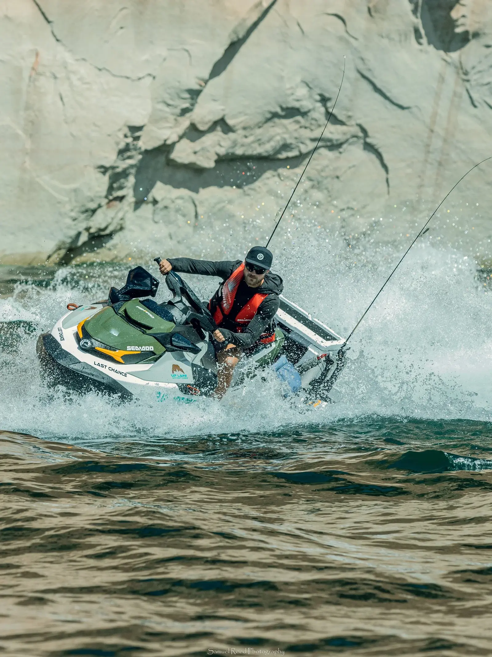 A man wearing sunglasses and a cap rides a jet ski on the water near rocky cliffs.