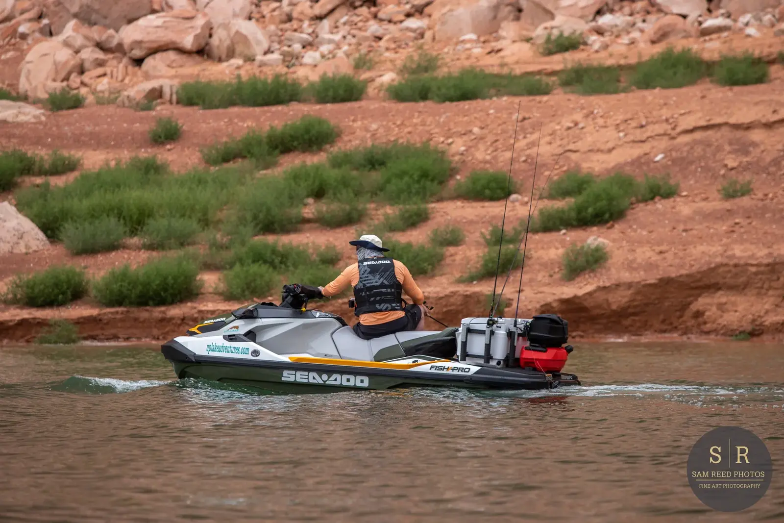 Person riding a Sea-Doo personal watercraft on a body of water with rocky, desert-like terrain and sparse green bushes in the background.
