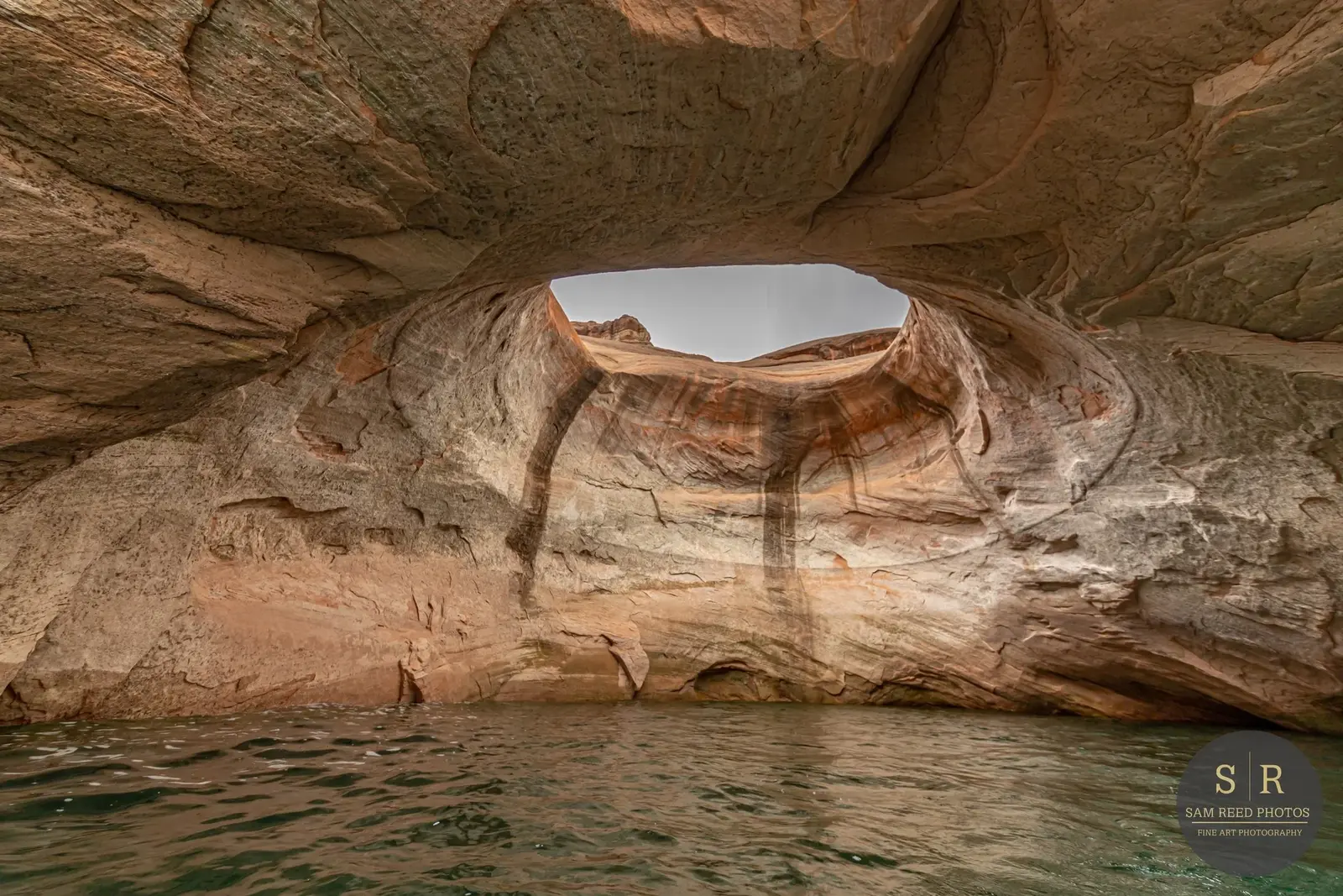 A natural rock formation with a hole in the ceiling, surrounded by tall, smooth, reddish-brown rock walls, above water in a canyon.