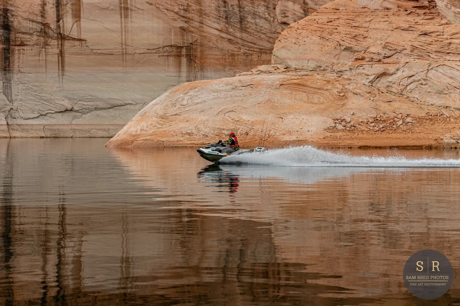 A person riding a jet ski on a calm body of water near large reddish-brown rocks or cliffs.