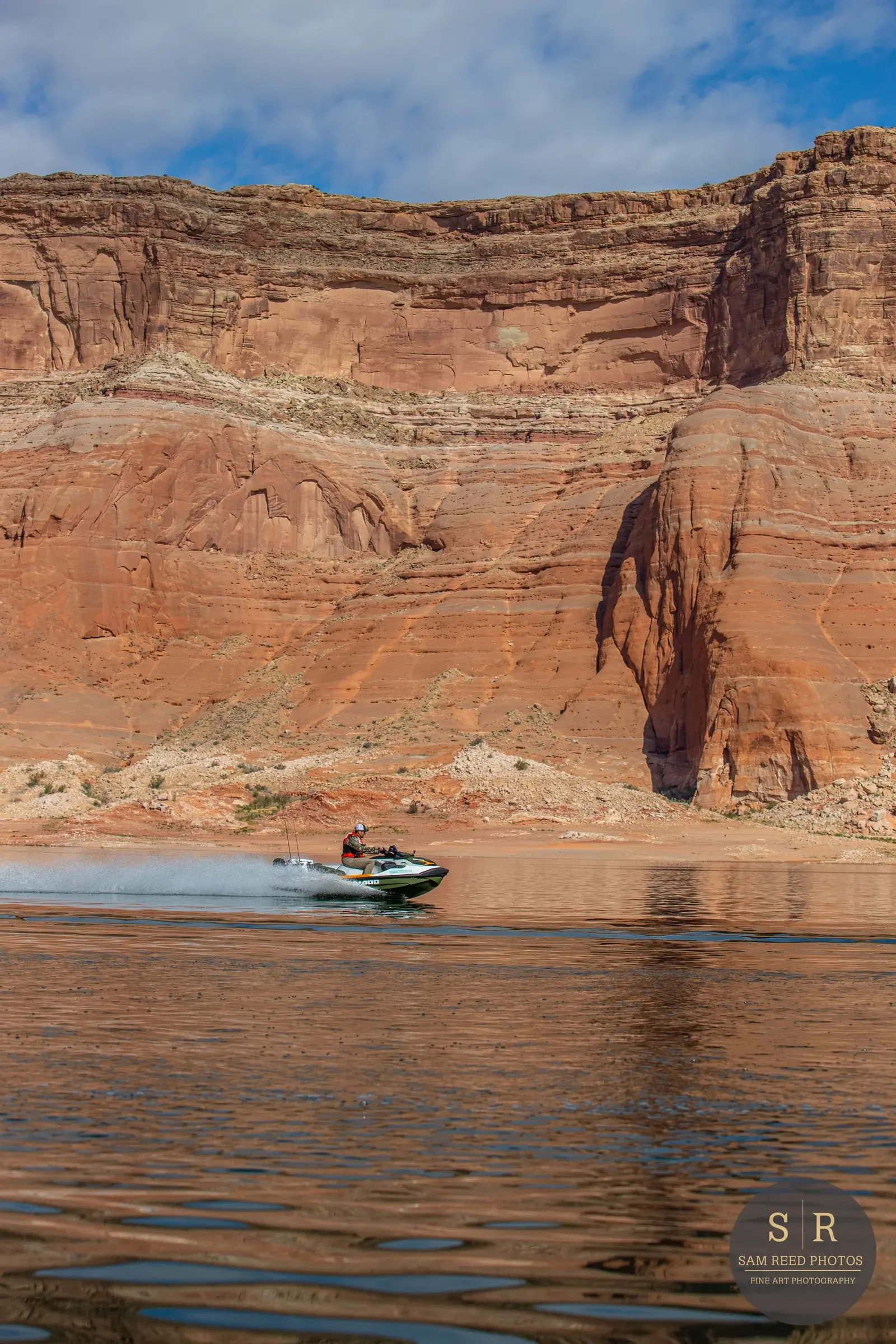 A person racing a jet ski on a lake with steep red rock formations and a partly cloudy sky in the background.