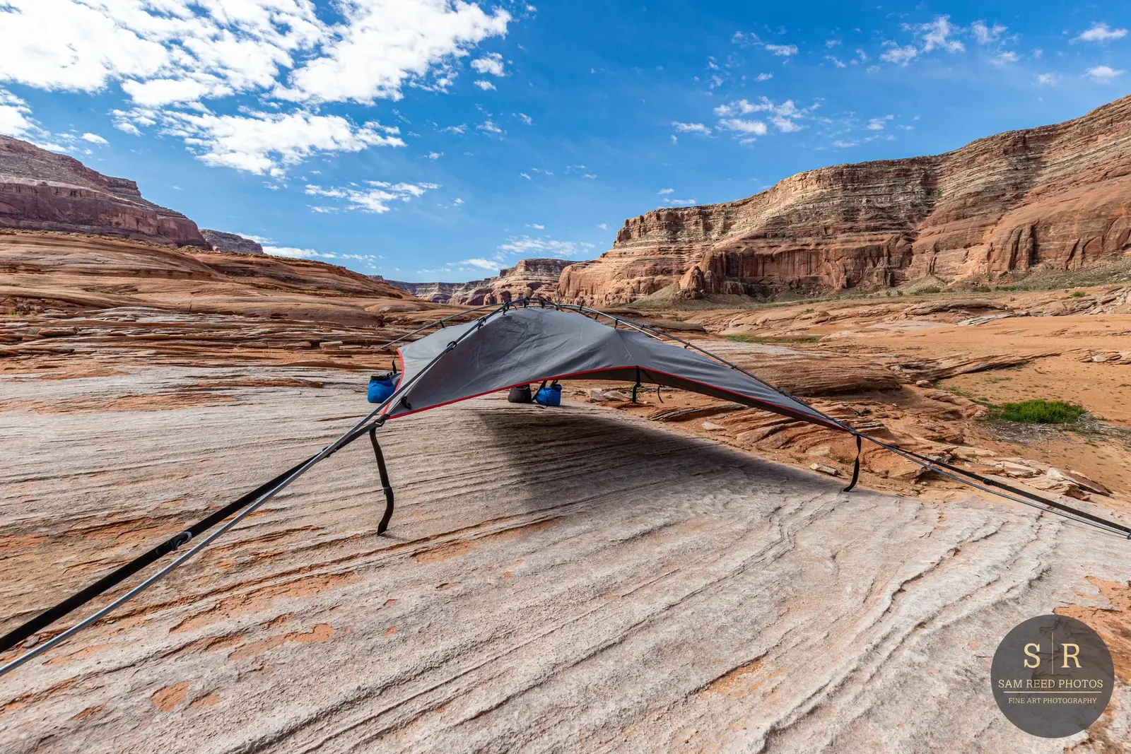A camping tent set up on a large, smooth rock formation in a desert landscape with red canyon walls in the background and a blue sky with scattered clouds above.