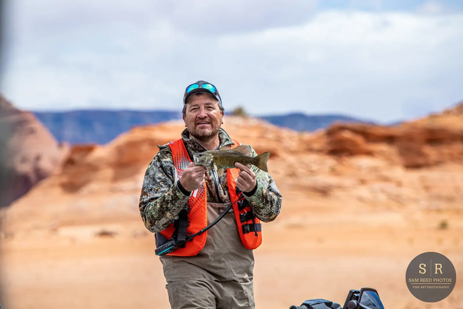 Man in camouflage jacket and orange life vest holding a fish outdoors in a desert landscape with rocky formations.