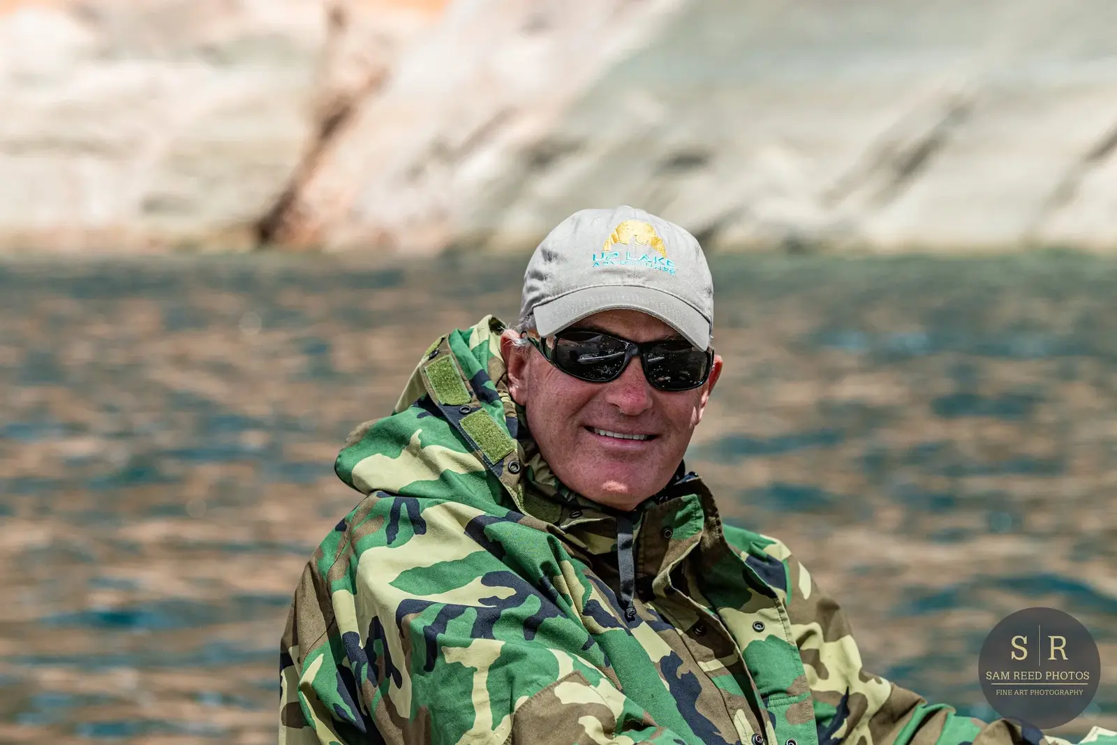 A man in camouflage jacket and sunglasses smiling outdoors near water with a rocky background.