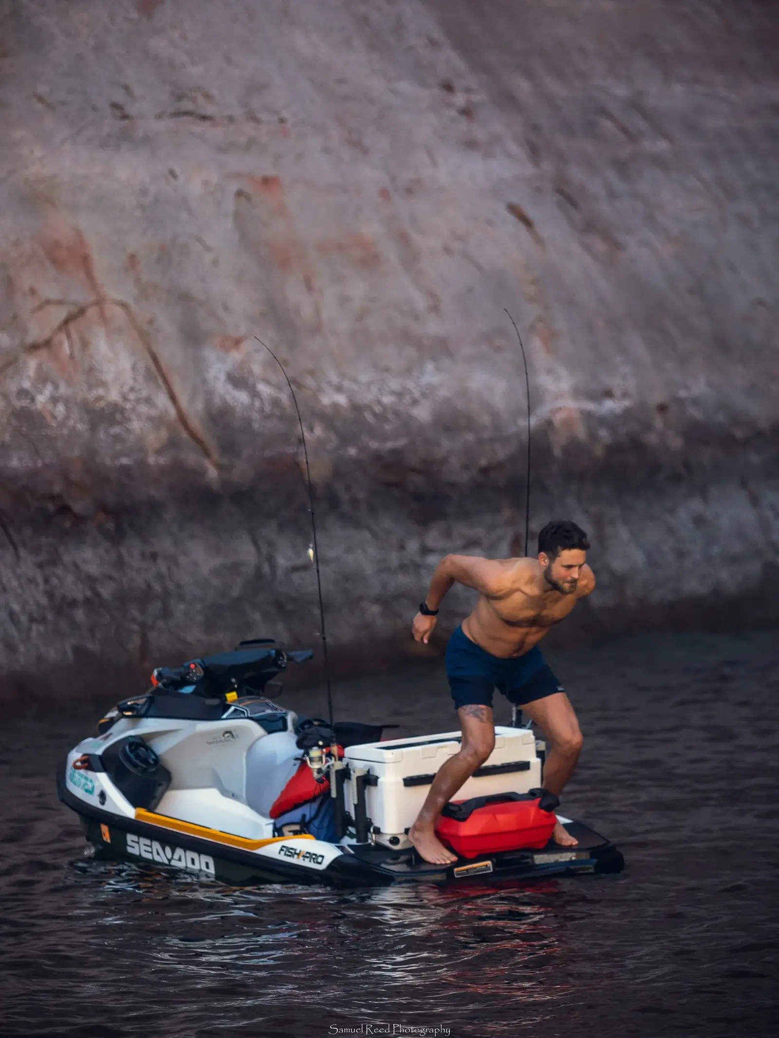 Man on an electric jet ski, shirtless and in black shorts, standing with knees bent, holding onto a box on the jet ski, near a rocky shoreline.
