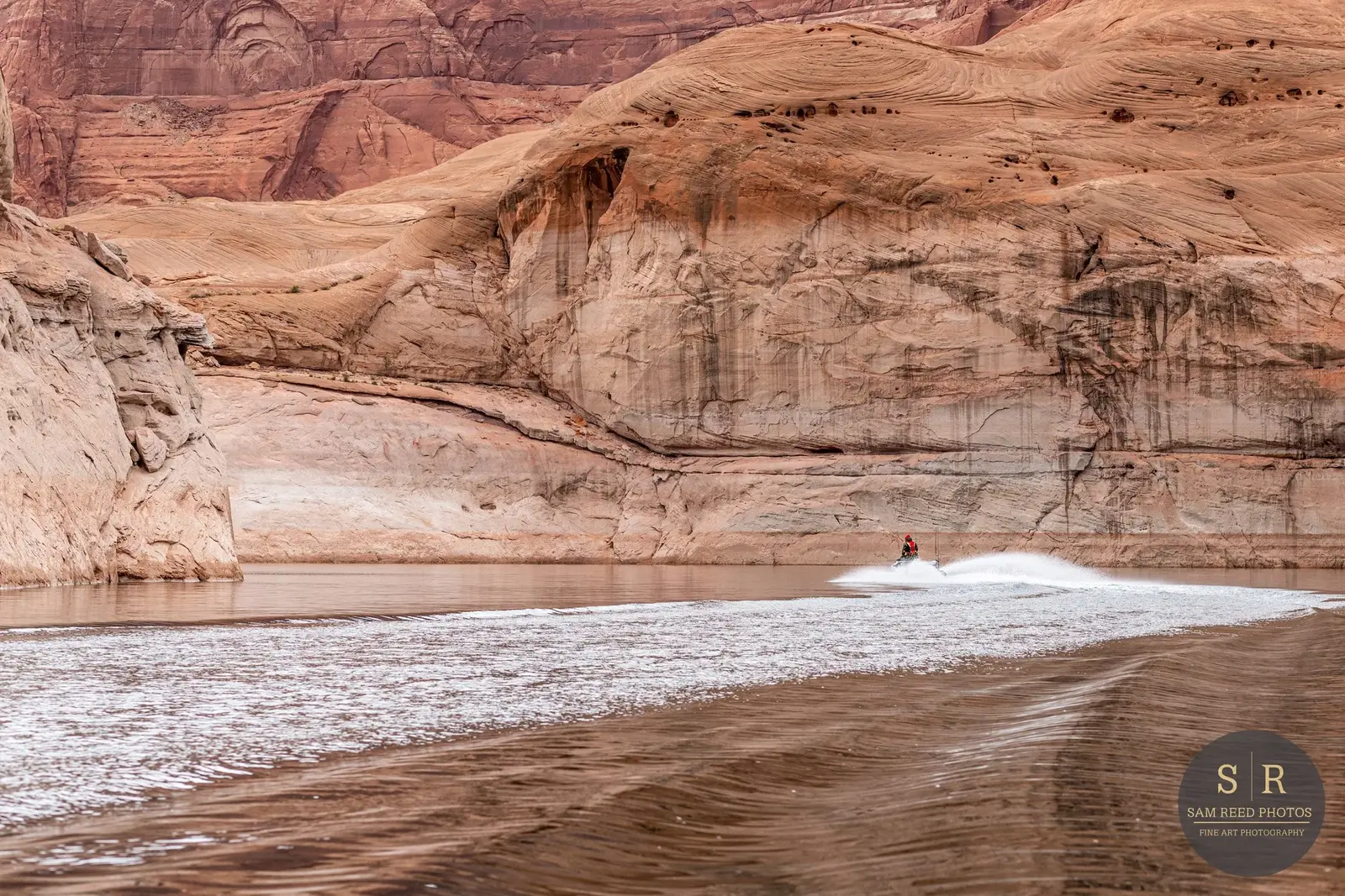 A boat speeding across a river surrounded by towering, reddish-brown canyon walls in a desert landscape.