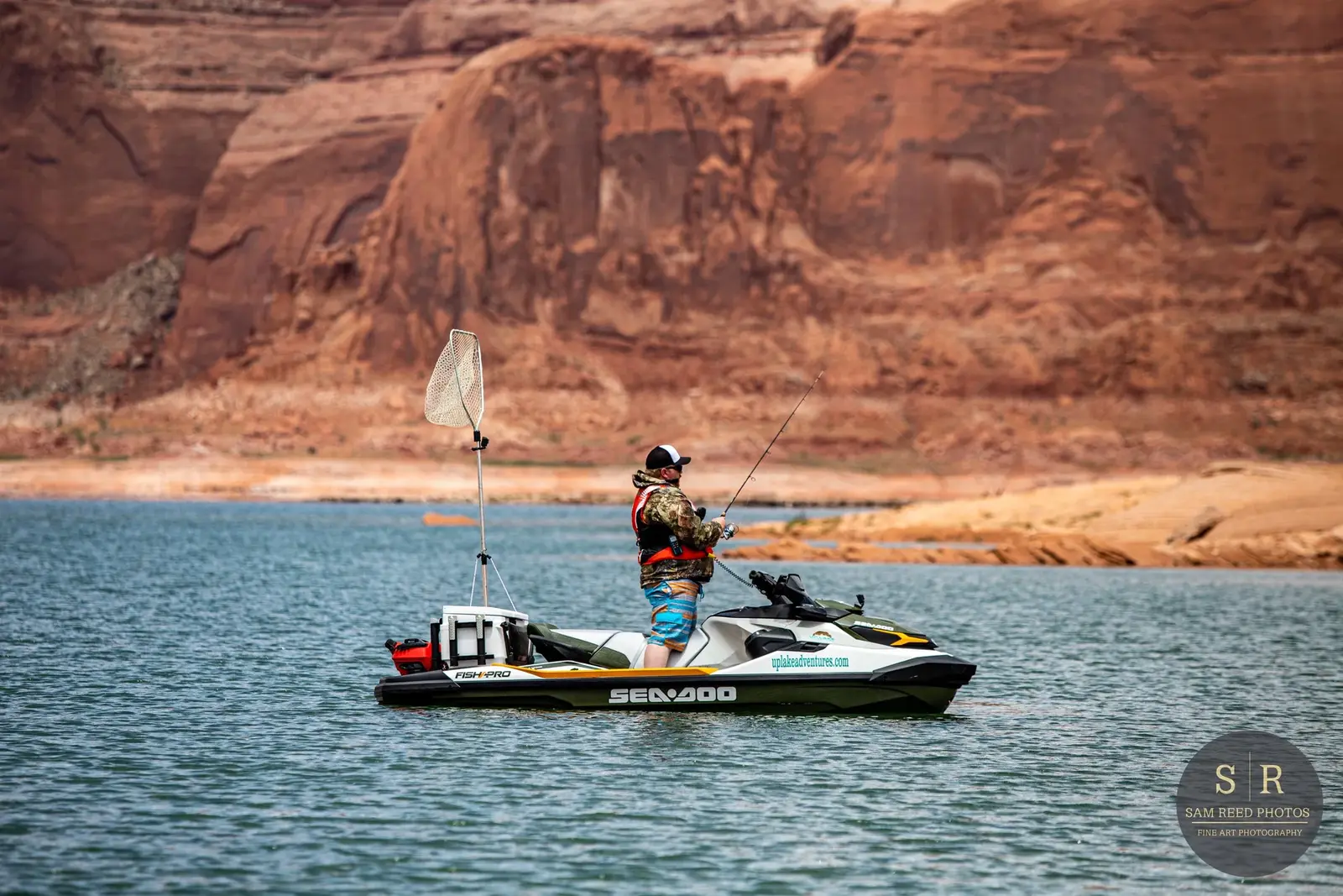Person fishing on a small motorboat on a lake with a desert landscape and red rock formations in the background.