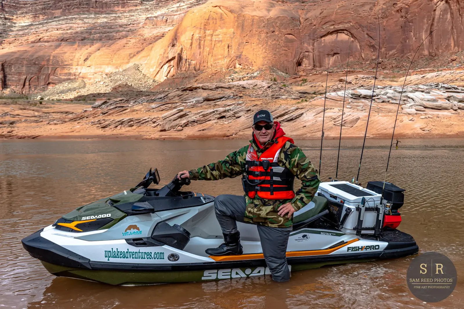 Man standing on a jet ski in a river with red rock cliffs in the background, wearing a red life jacket, camouflage jacket, black hat and sunglasses.