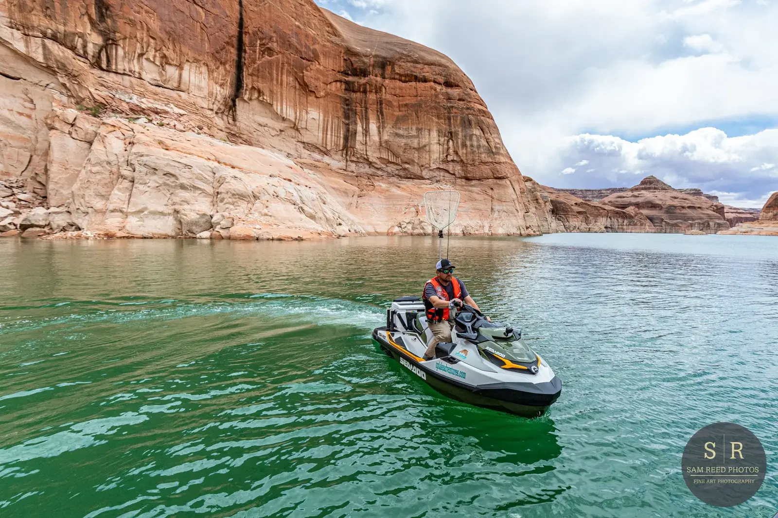 Man riding a jet ski on a body of water near red rock cliffs with cloudy sky in the background.