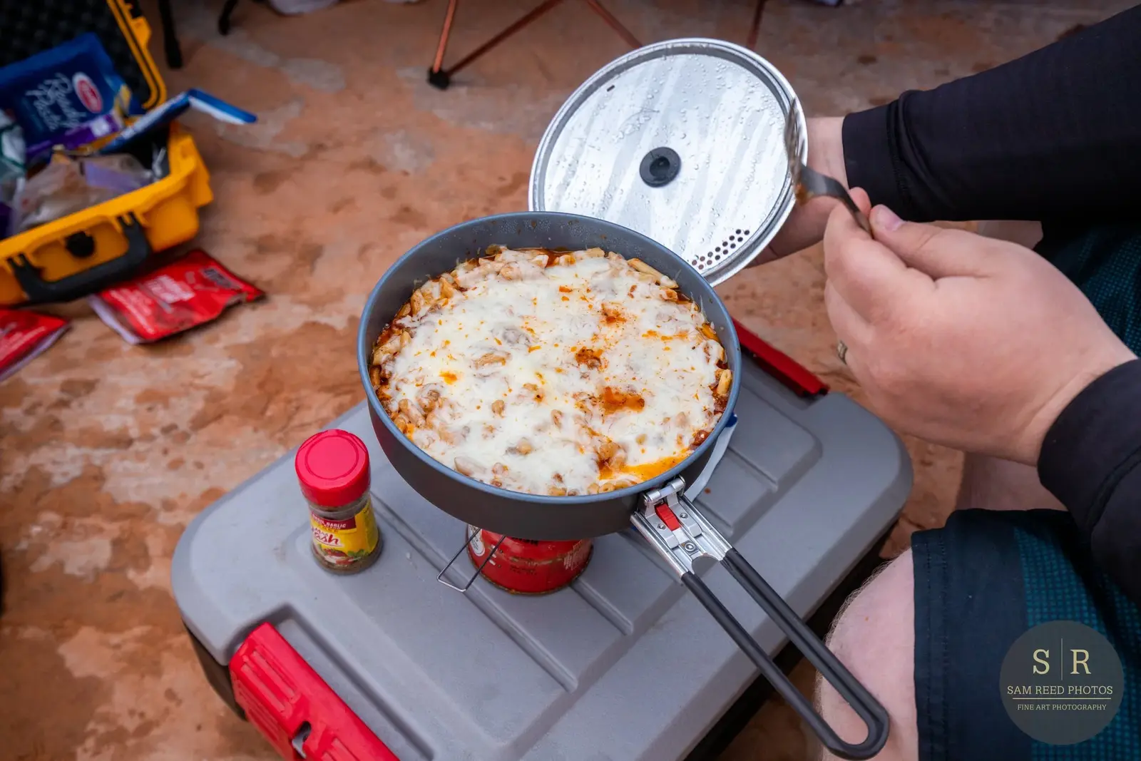 Person holding a pot of baked pasta or lasagna over a portable stove during a camping trip.