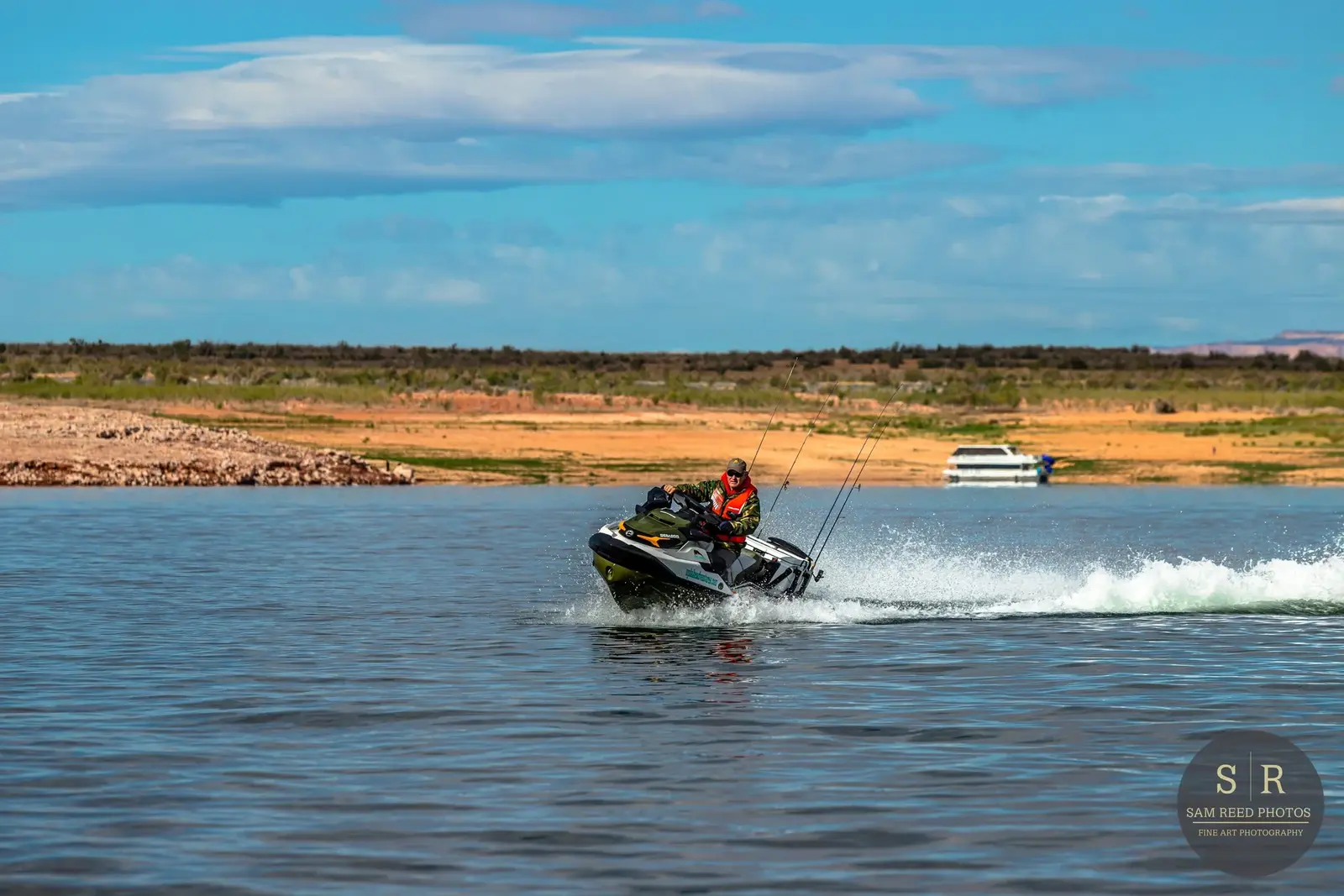 A person riding a jet ski on a body of water with a distant boat and a dry, rocky shoreline under a partly cloudy sky.