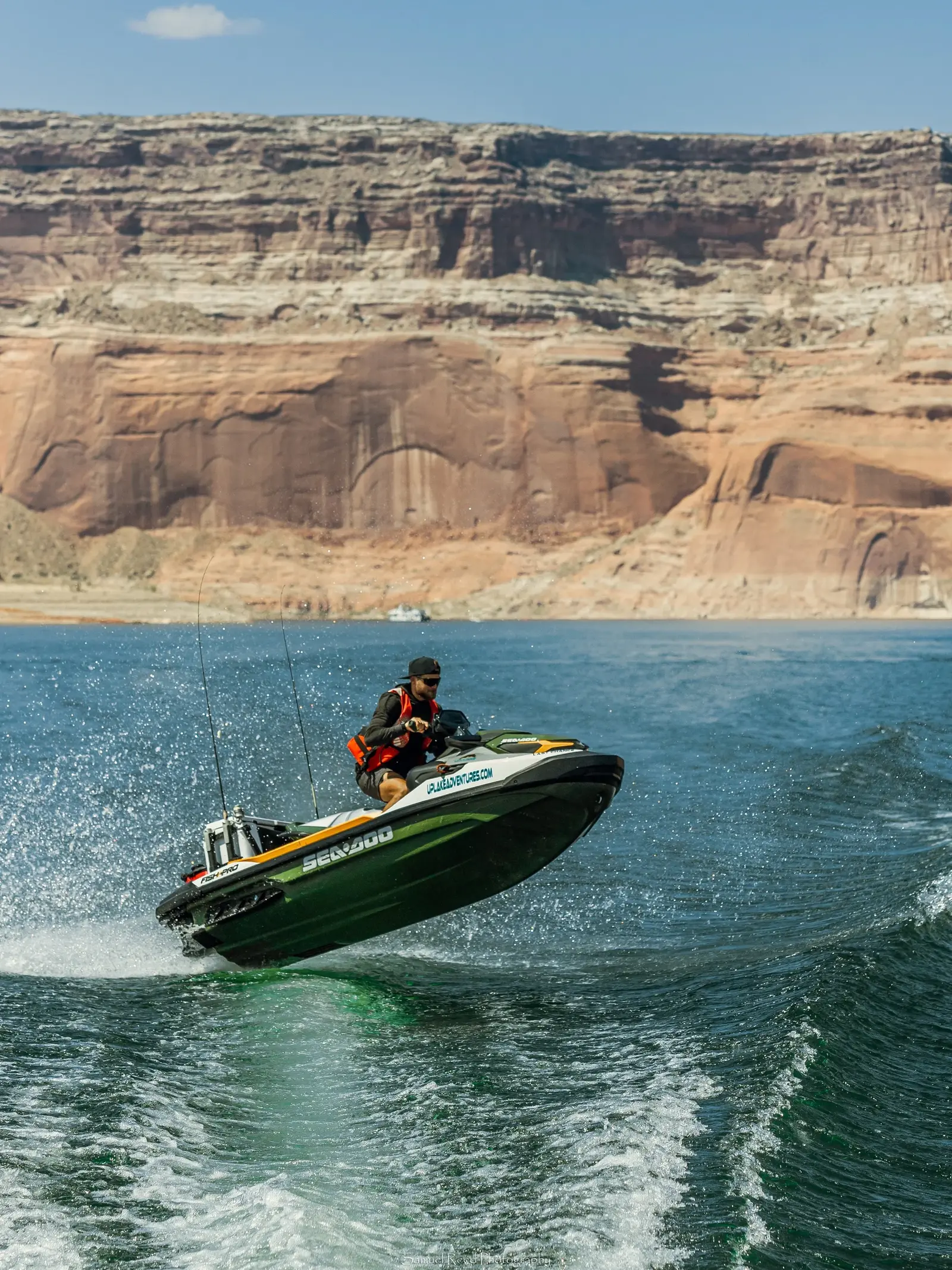 A person riding a green and black Seadoo jet ski on a large body of water with red rock cliffs in the background.