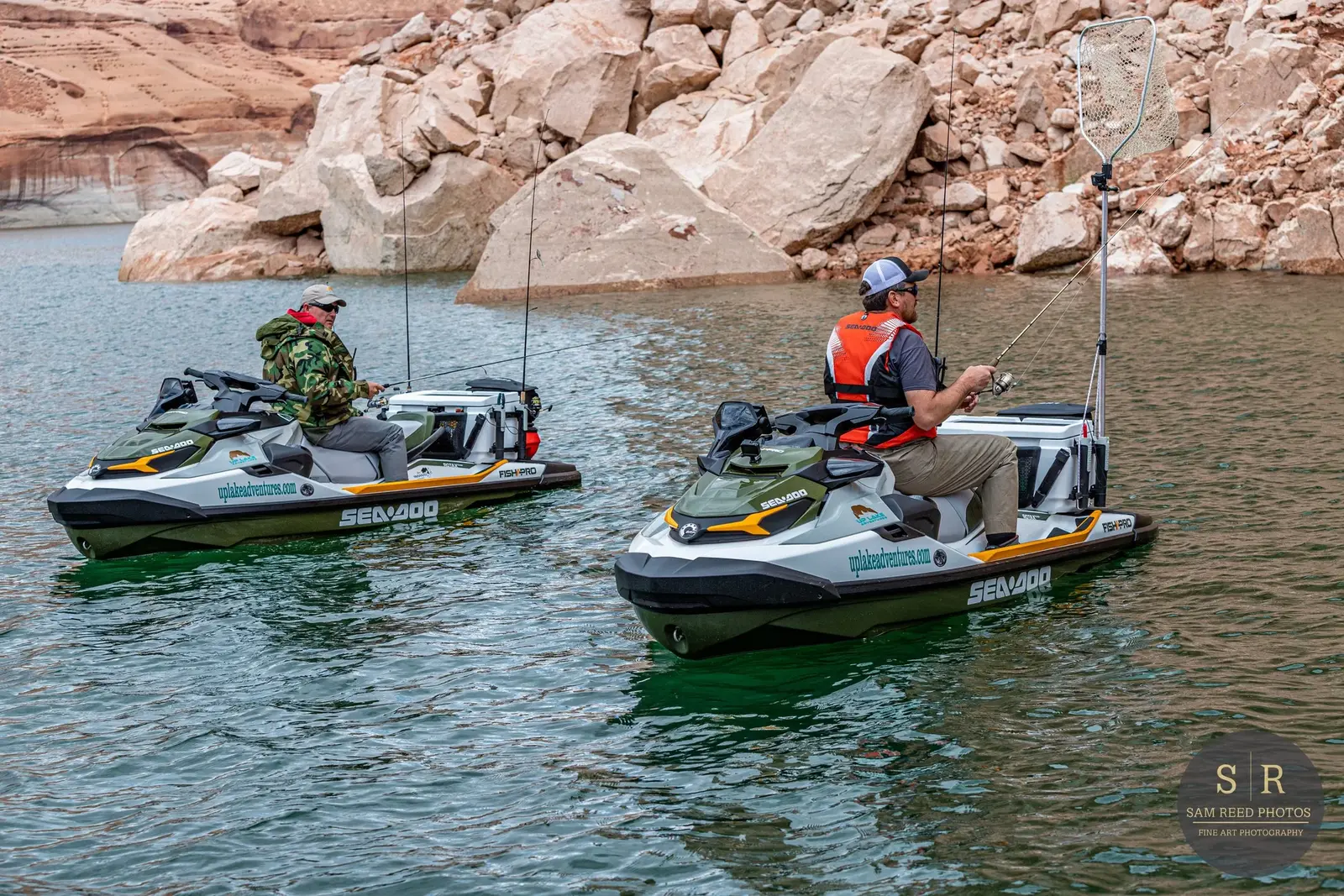 Two people fishing from jet skis on a lake with rocky cliffs in the background.
