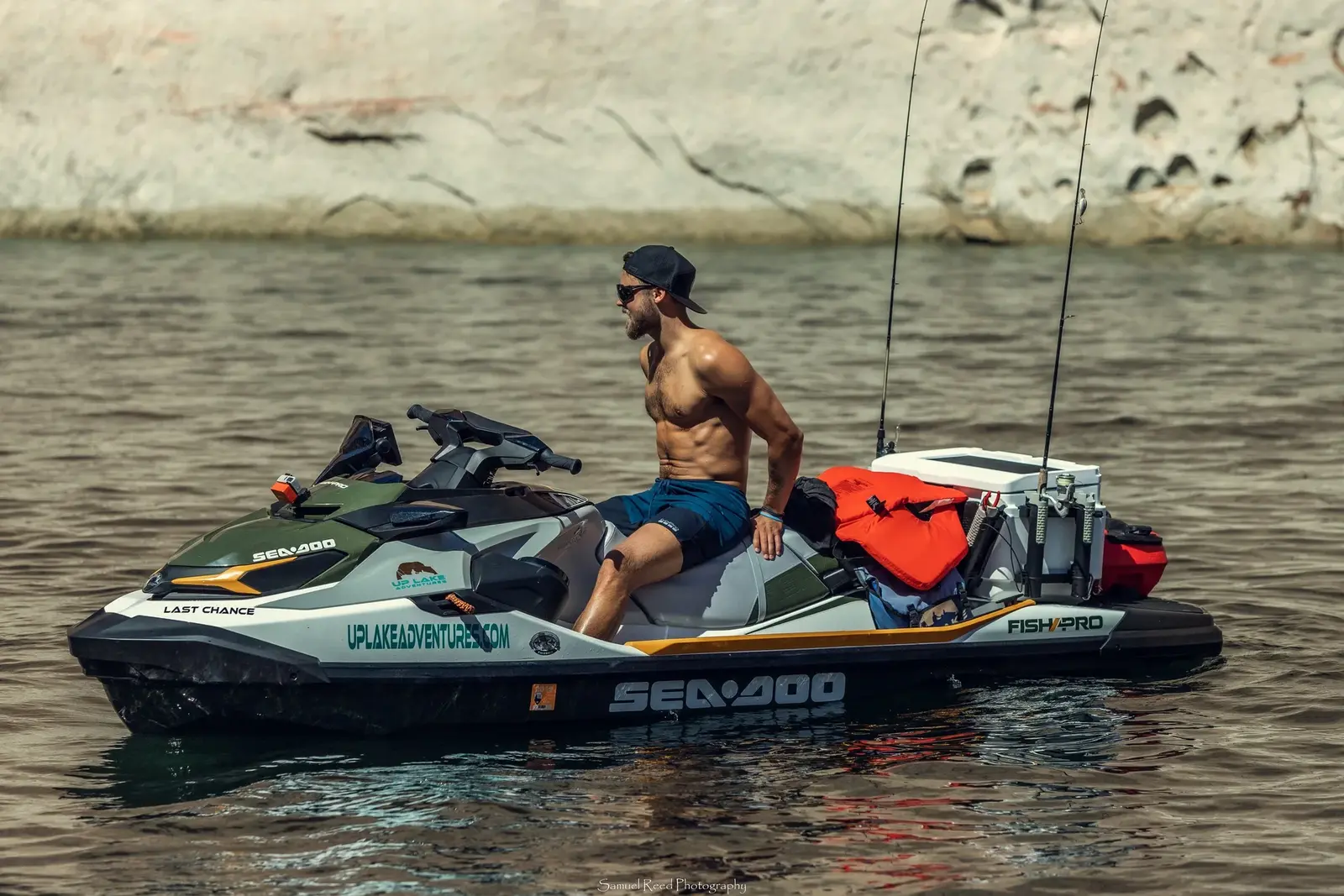 A shirtless man wearing sunglasses, a backwards cap, and shorts driving a jet ski on water near a rocky shoreline.