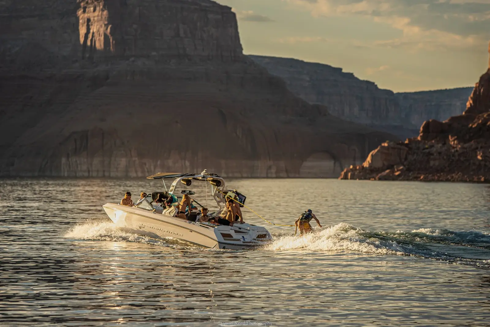 A group of people on a motorboat being towed by a person on a jet ski on a large body of water with canyon cliffs in the background at sunset.