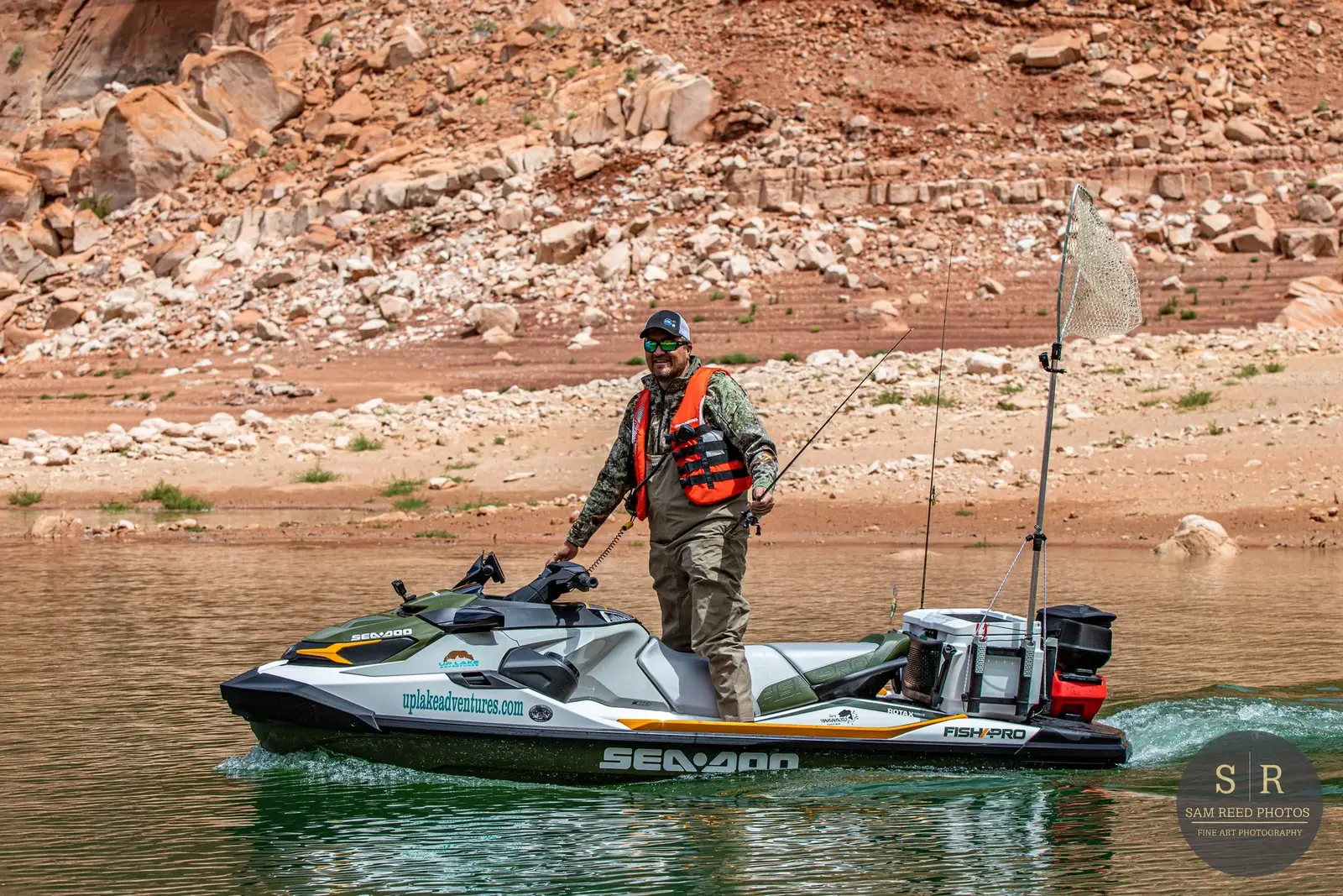A man on a jet ski fishing in a body of water with a rocky, reddish landscape in the background.