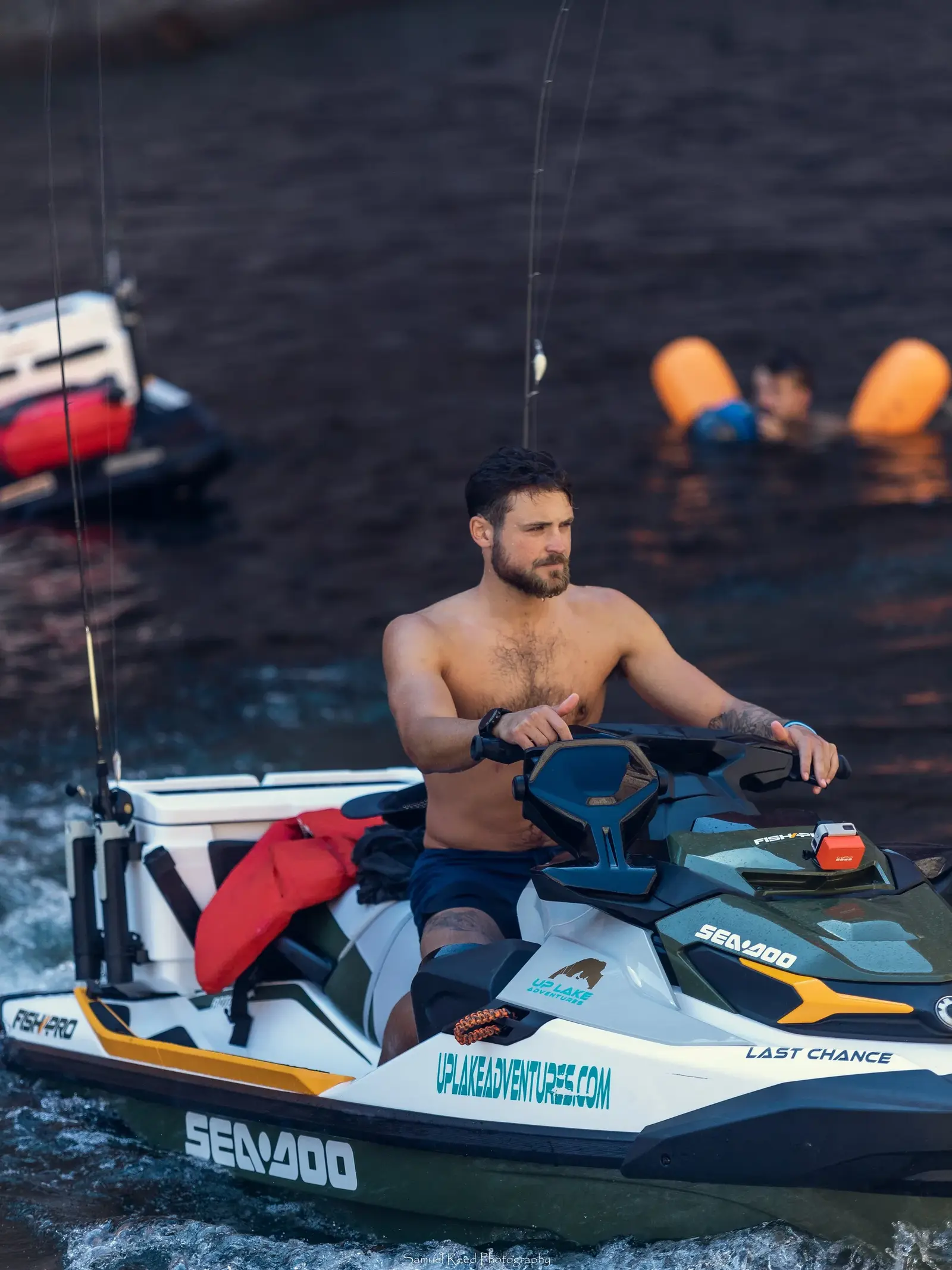 A shirtless man is riding a Sea-Doo jet ski on the water with another person swimming nearby, using orange flotation devices.