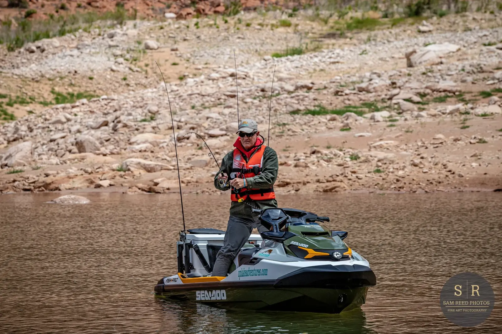 A man fishing on a Sea-Doo jet ski in a calm body of water with rocky shore and sparse green vegetation in the background.