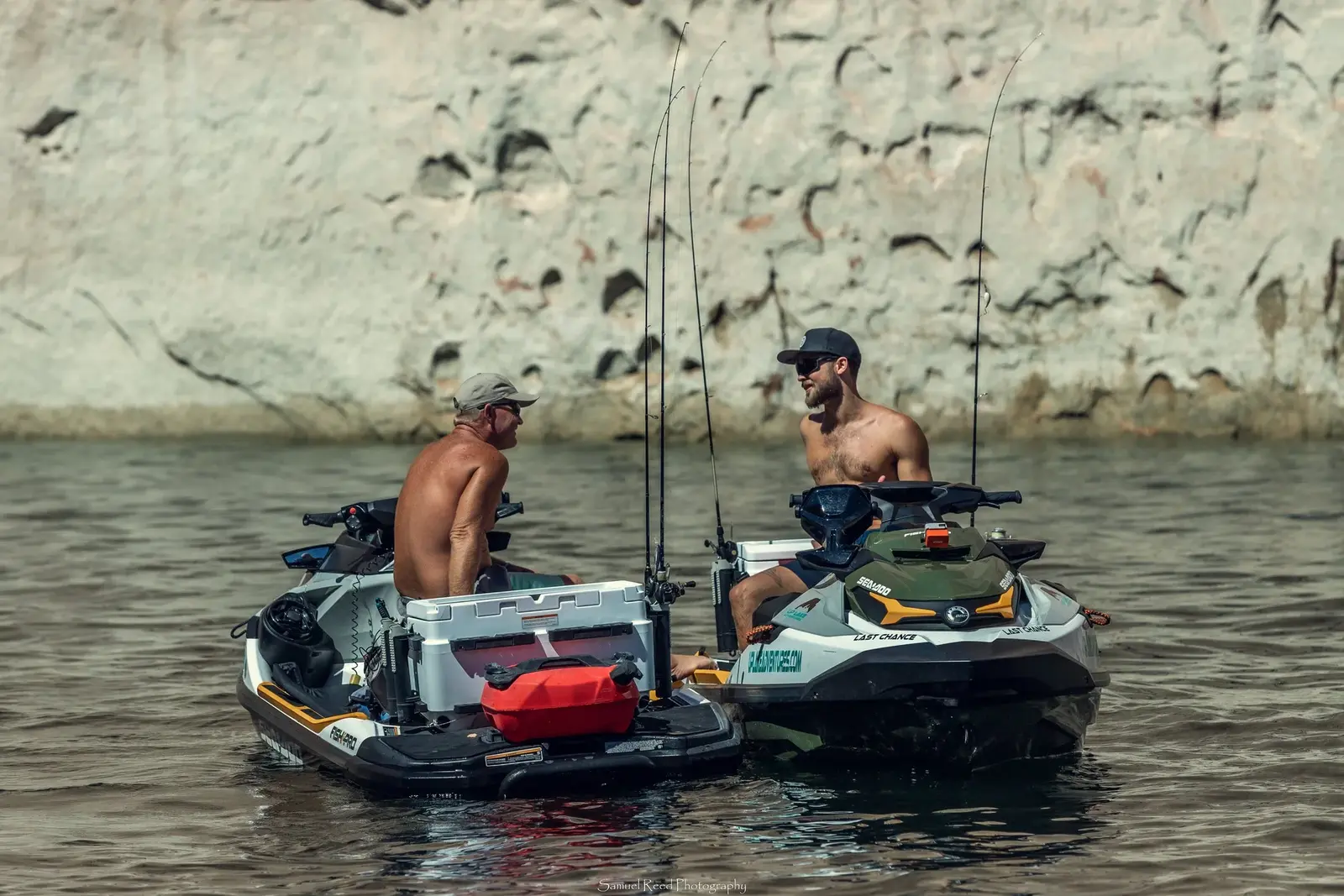 Two shirtless men with caps and sunglasses sitting on personal watercrafts (jet skis) having a conversation near a rocky shoreline.