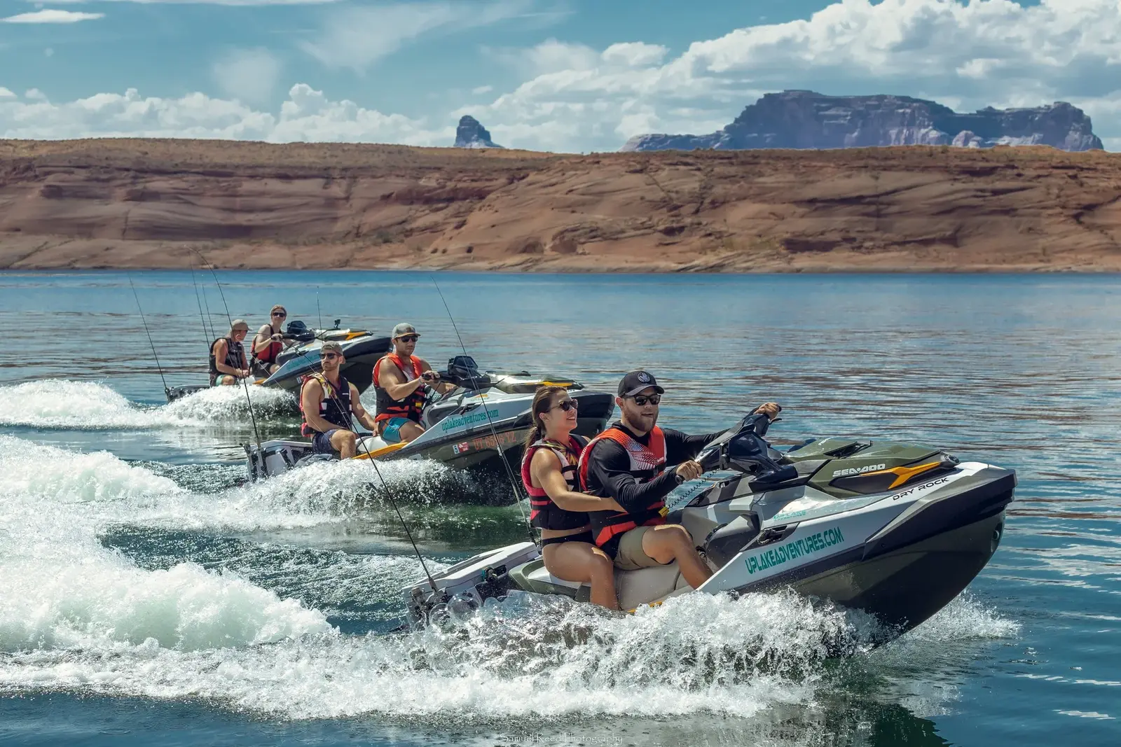 Six people riding on jet skis in a lake with rocky cliffs and a cloudy sky in the background.