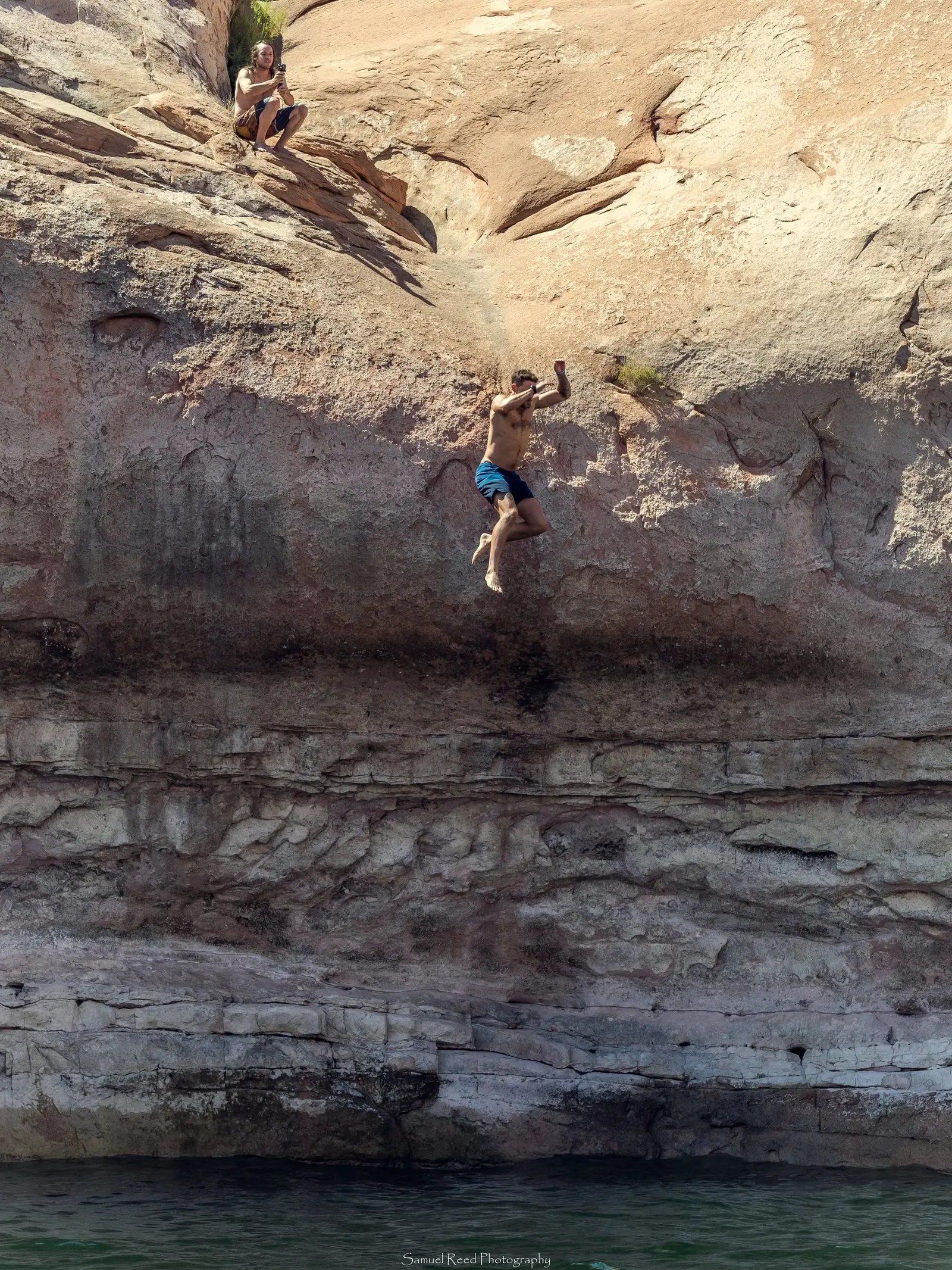 A shirtless man in blue shorts is jumping off a rock ledge into water below, while a young woman is sitting on the rocks above taking a photo or video with her phone.