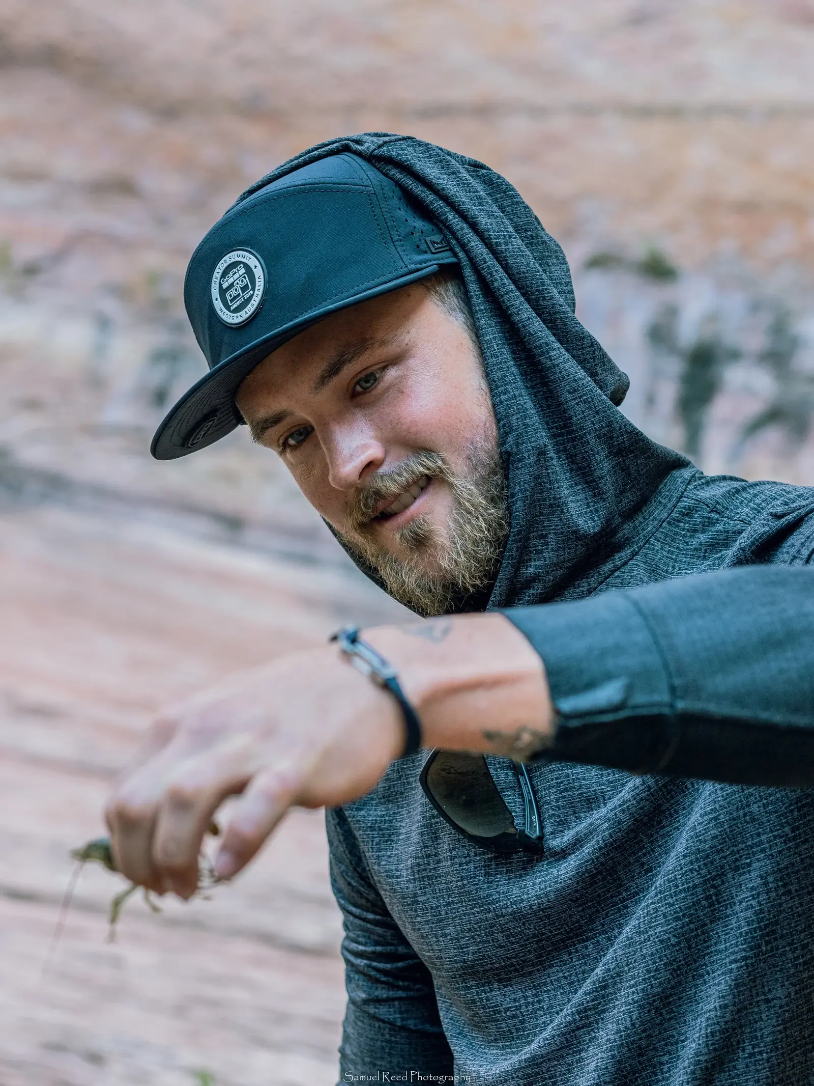 A man with a beard wearing a dark cap, hooded jacket, and sunglasses displays a small insect on his finger outdoors against a blurred rock background.