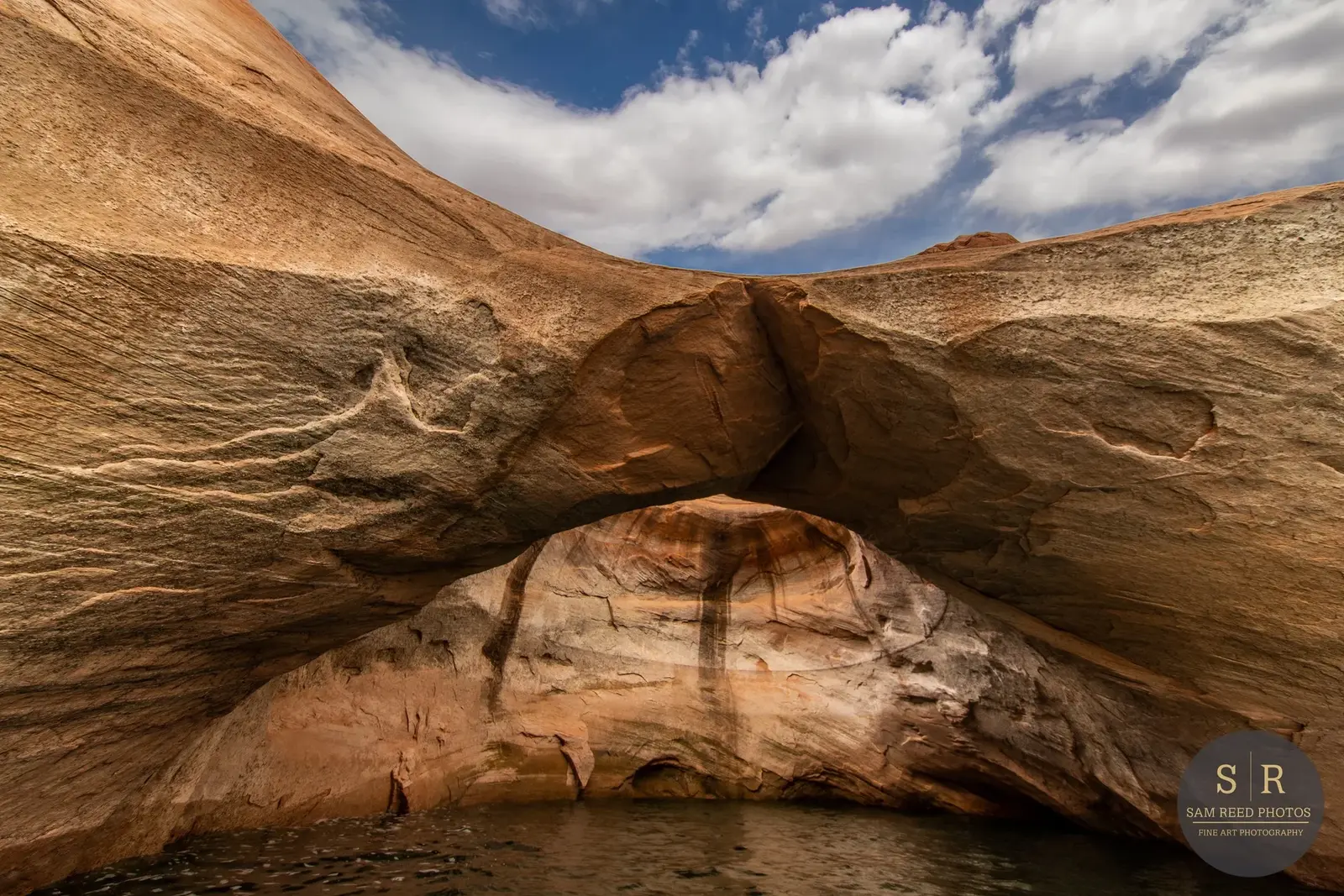 Natural rock formation with an arch haloed by blue sky and white clouds. The lower part of the formation is reflected in water.