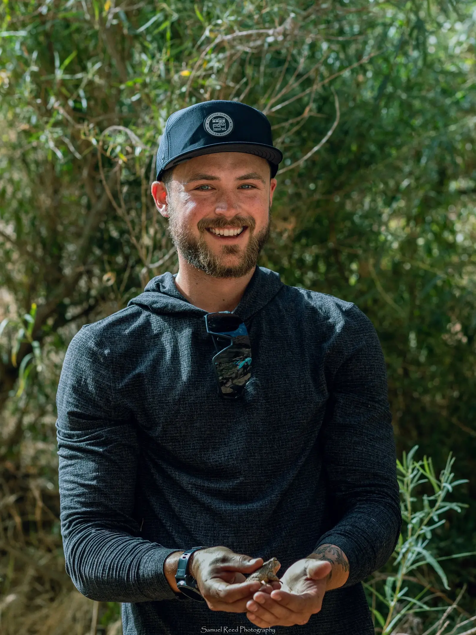 A smiling man with a beard and sunglasses hanging from his hoodie, holding a small animal or object, standing outdoors with green foliage in the background.