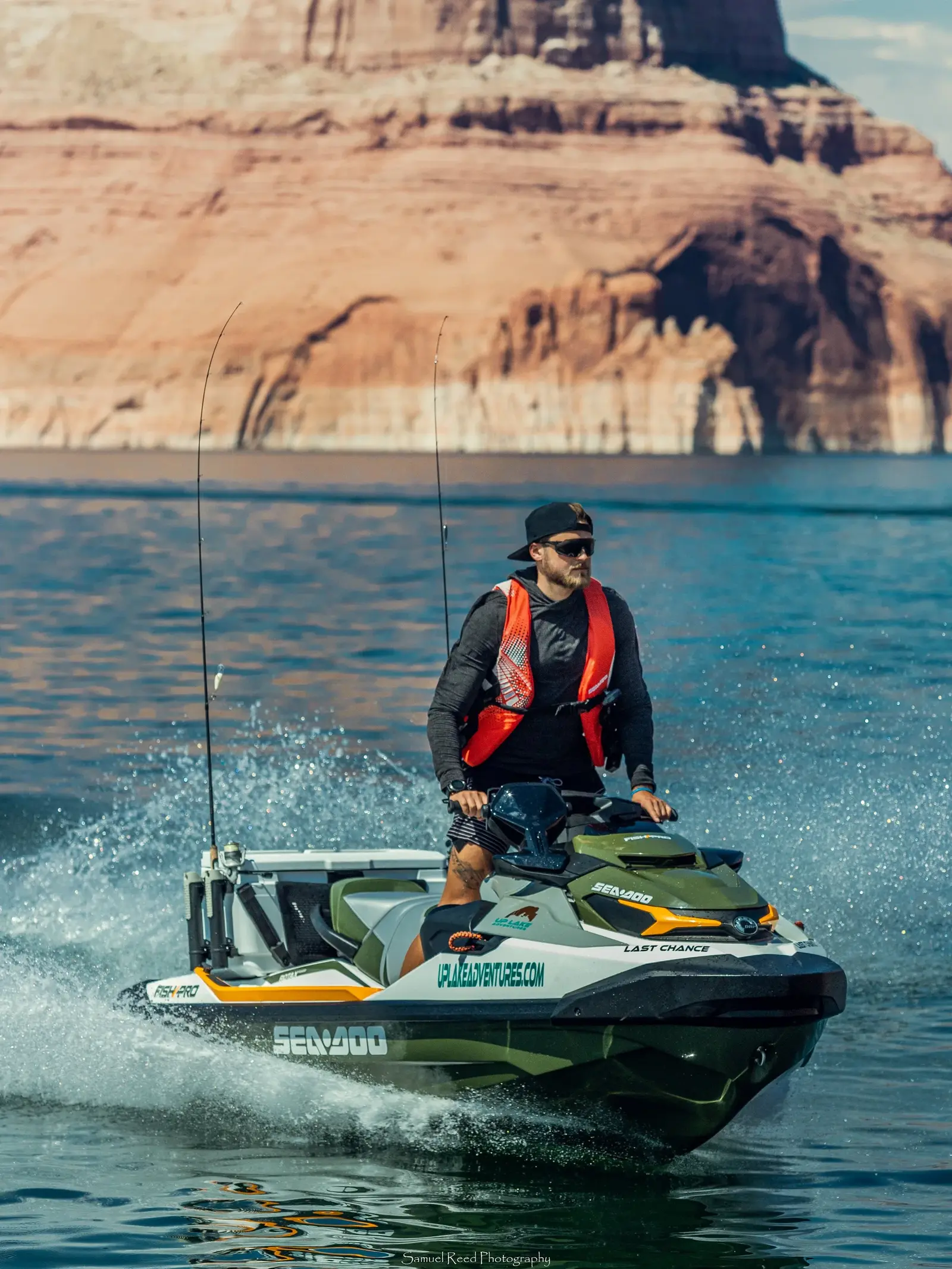 A man riding a green and white Seadoo jet ski on a lake with a large red rock formation in the background.