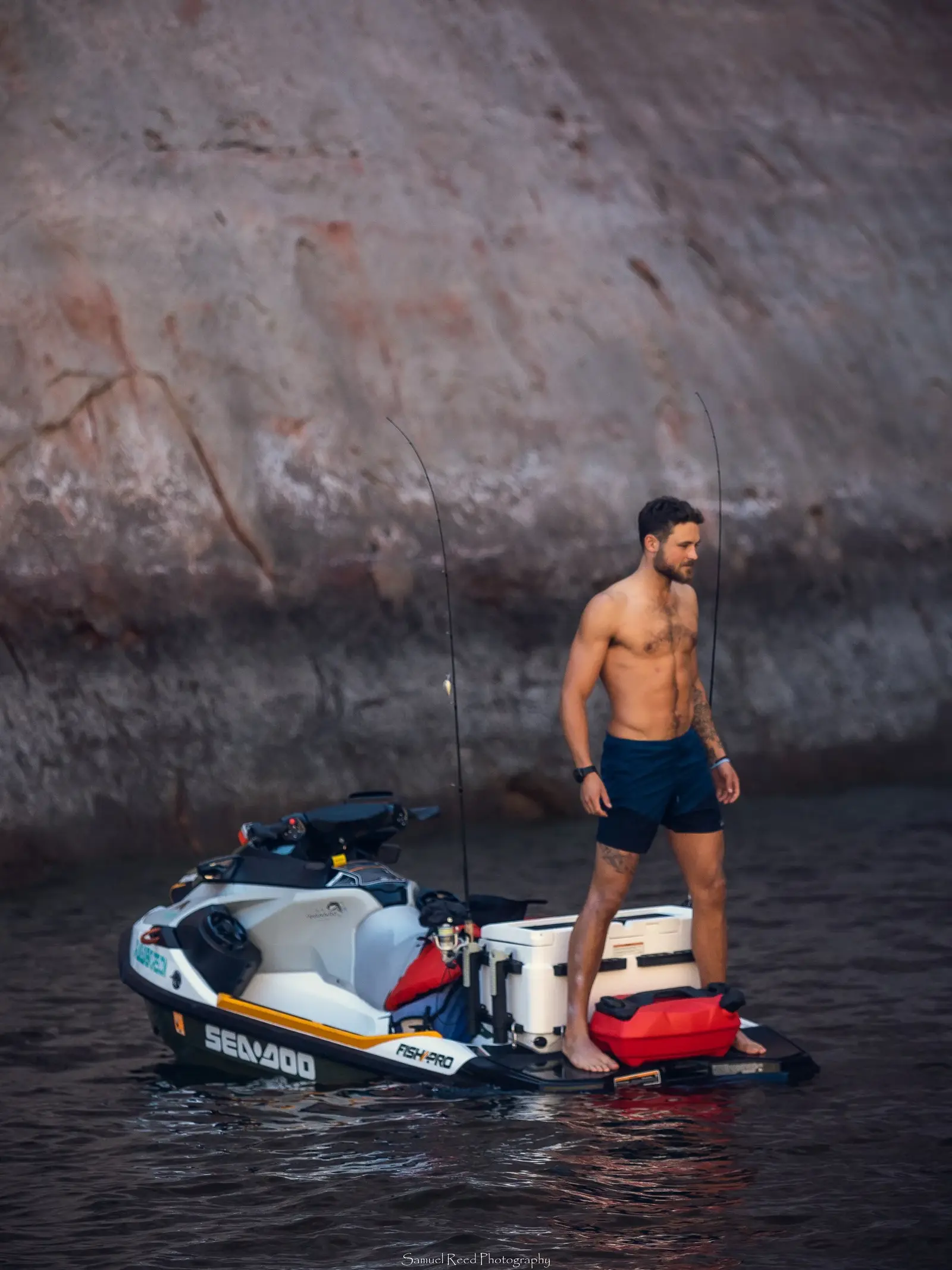 A shirtless man with tattoos standing on a jet ski near a rocky shoreline.