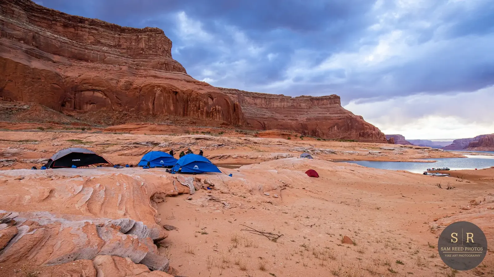 Campsite with multiple tents near a lake in a desert landscape. Rocky cliffs and a cloudy sky are visible in the background.