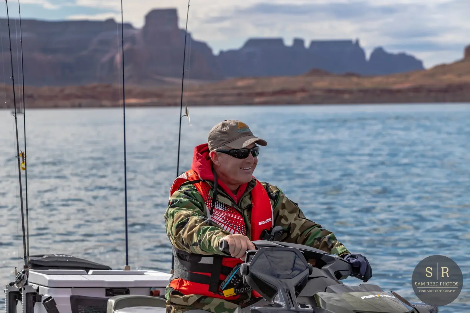 A man driving a boat on a lake with mountains in the background, wearing sunglasses, a camouflage jacket, and a red life jacket.