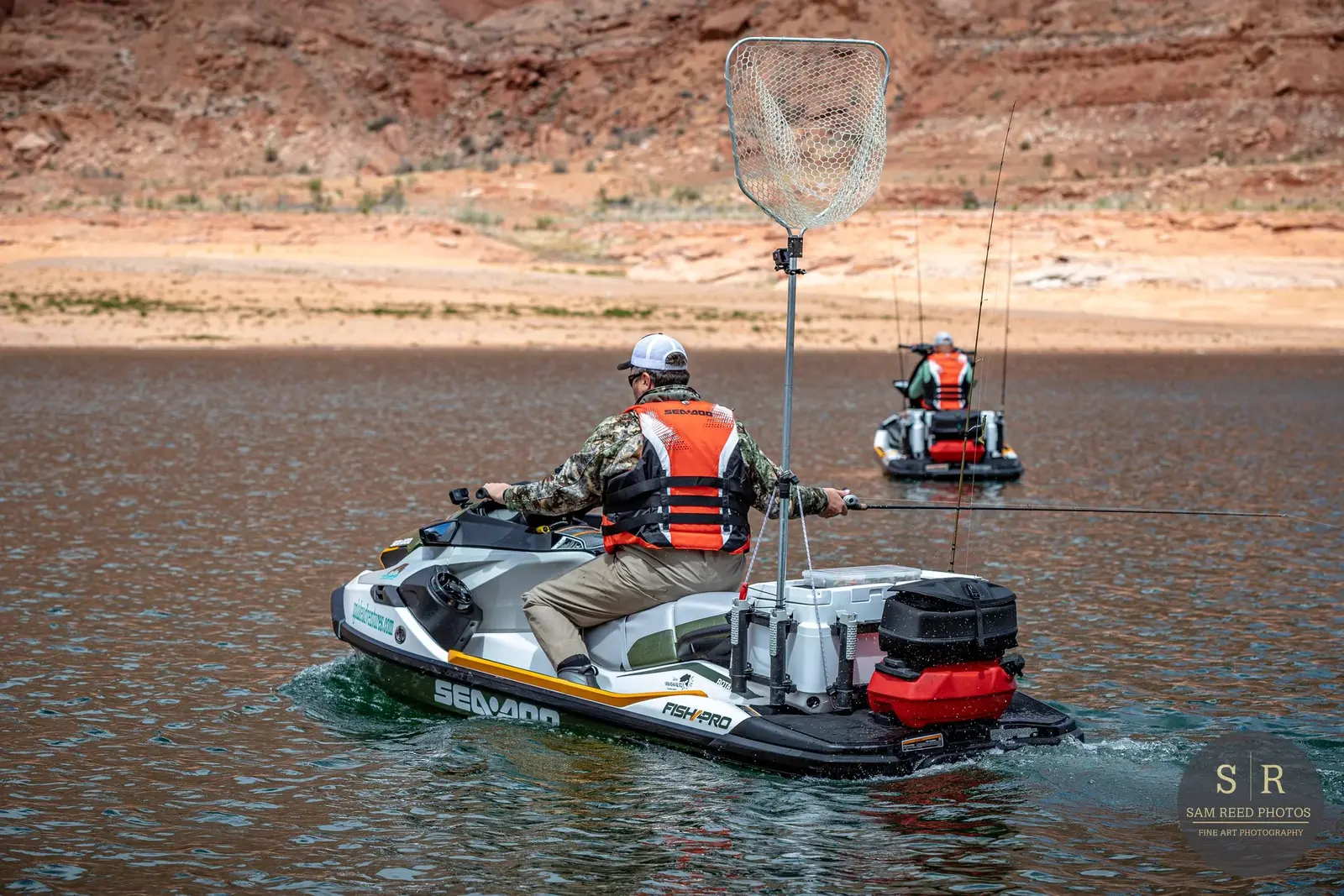 Two fishermen on personal watercraft fishing in a large lake with desert landscape and red rock formations in the background.
