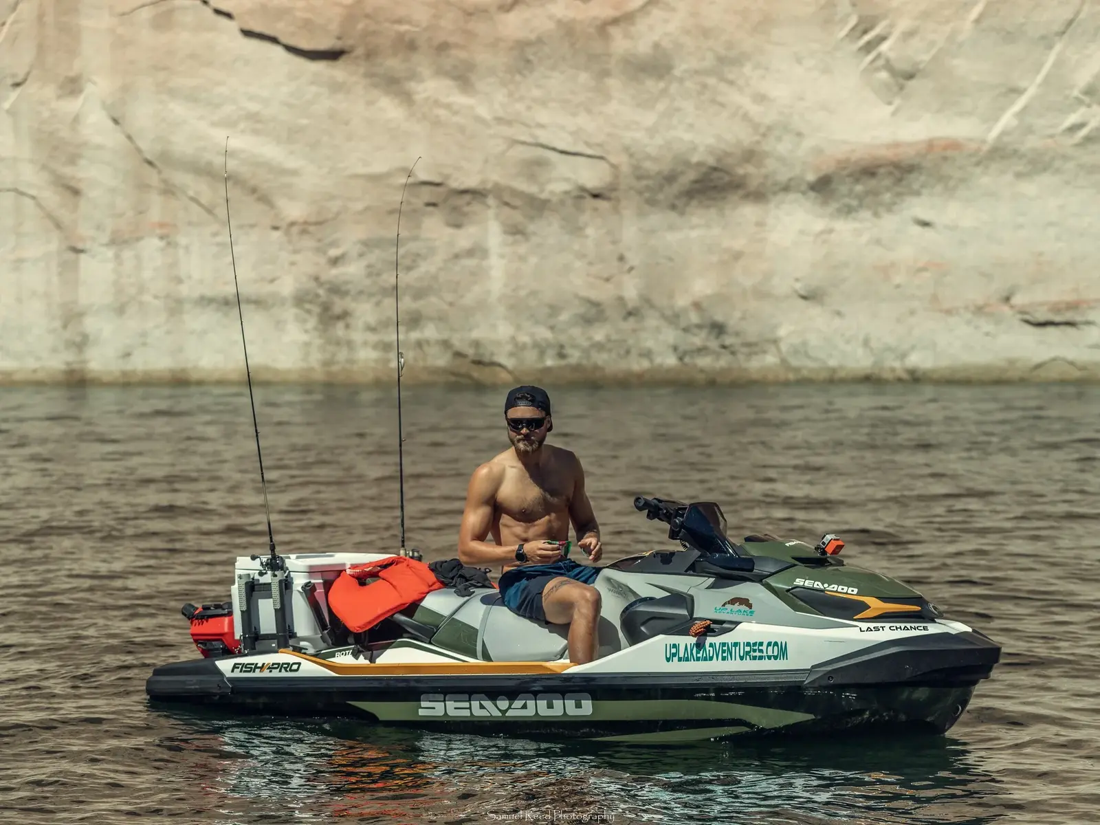 A man in sunglasses and a backwards cap is sitting shirtless on a Sea-Doo jet ski on a body of water, with a rocky cliff face in the background.