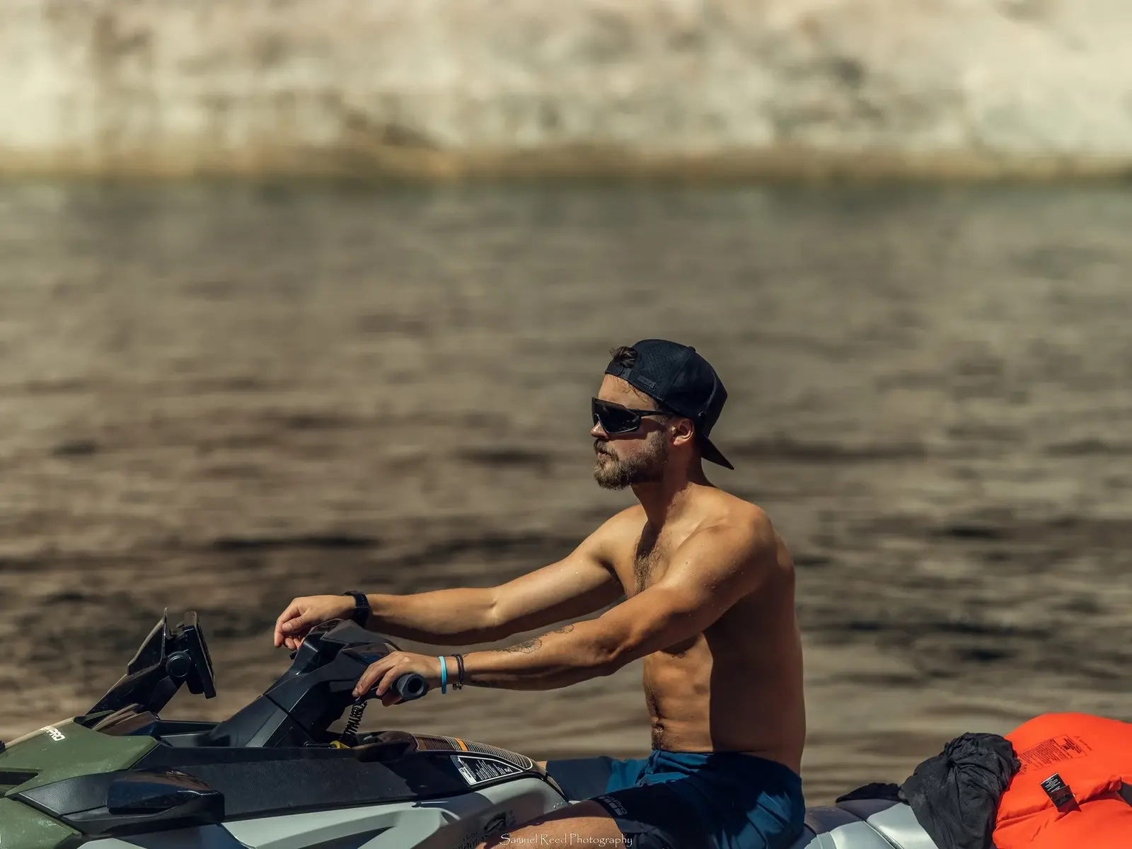 A shirtless man wearing sunglasses and a backward cap rides a jet ski on water, with a blurred background of a beach or rocky shoreline. An orange life jacket and a gray cloth are on the jet ski behind him.