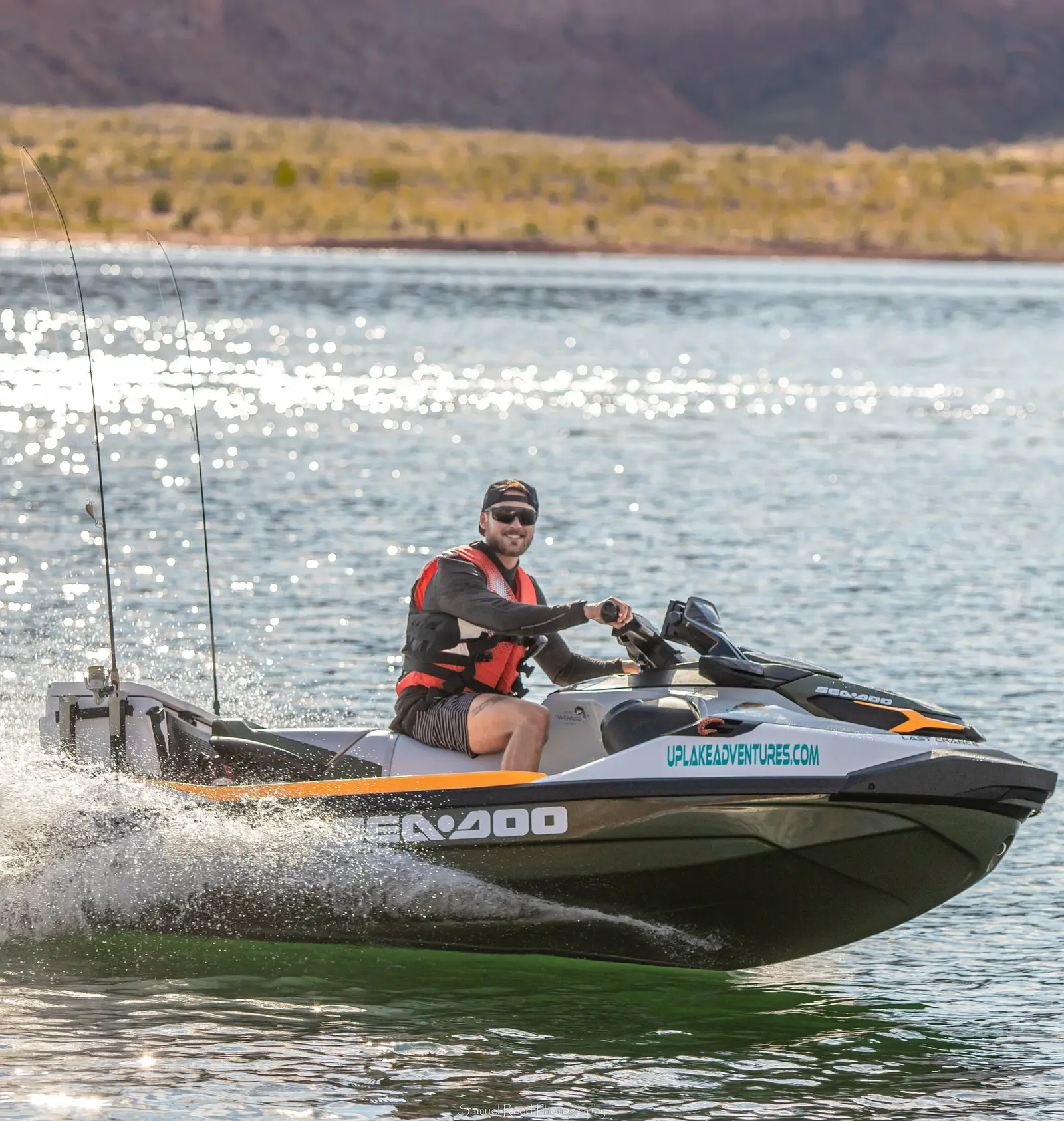 A man riding a Sea-Doo jet ski on a lake, smiling, with mountains and a shoreline in the background.