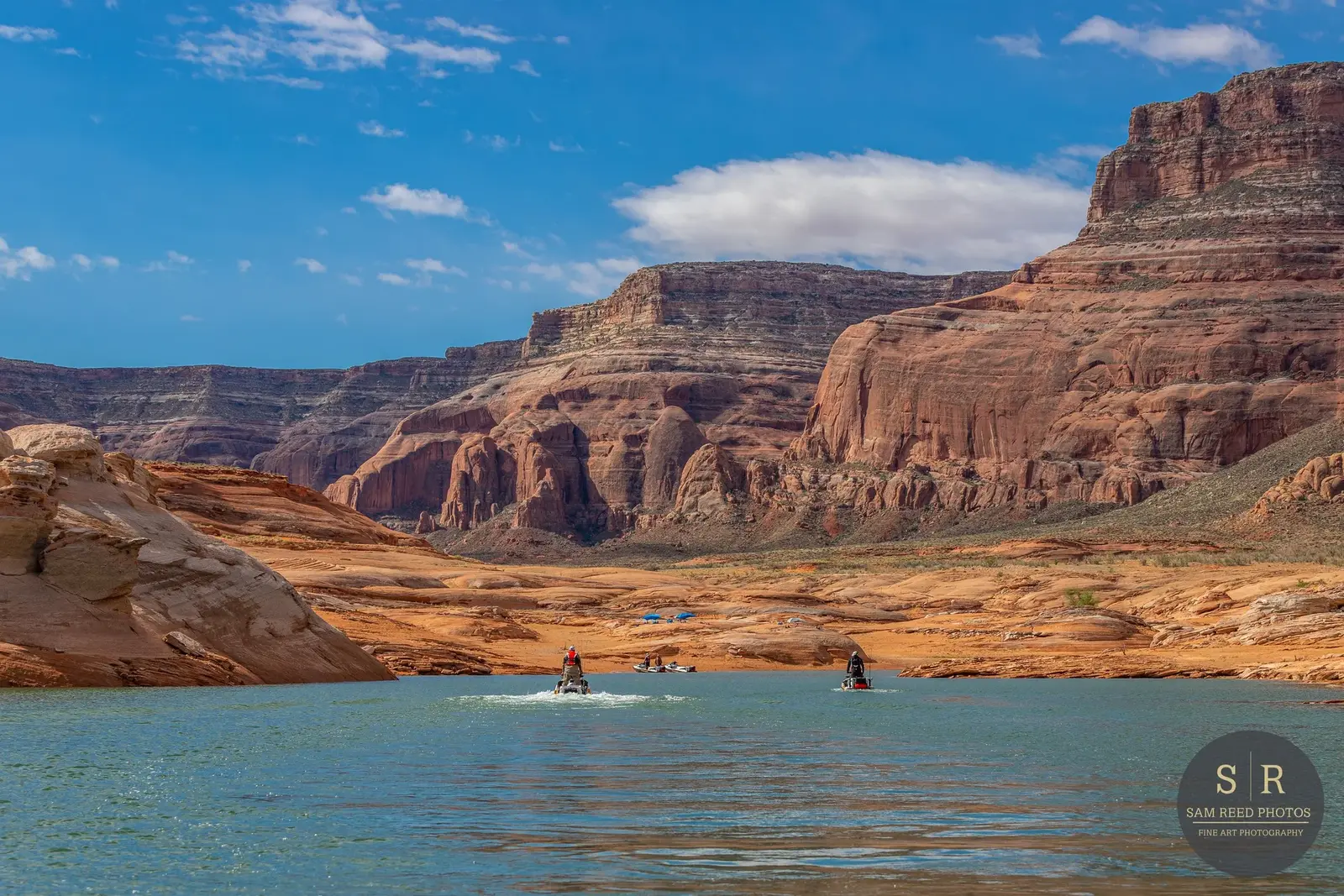 People kayaking on a river surrounded by red rock formations and mountains under a partly cloudy sky.