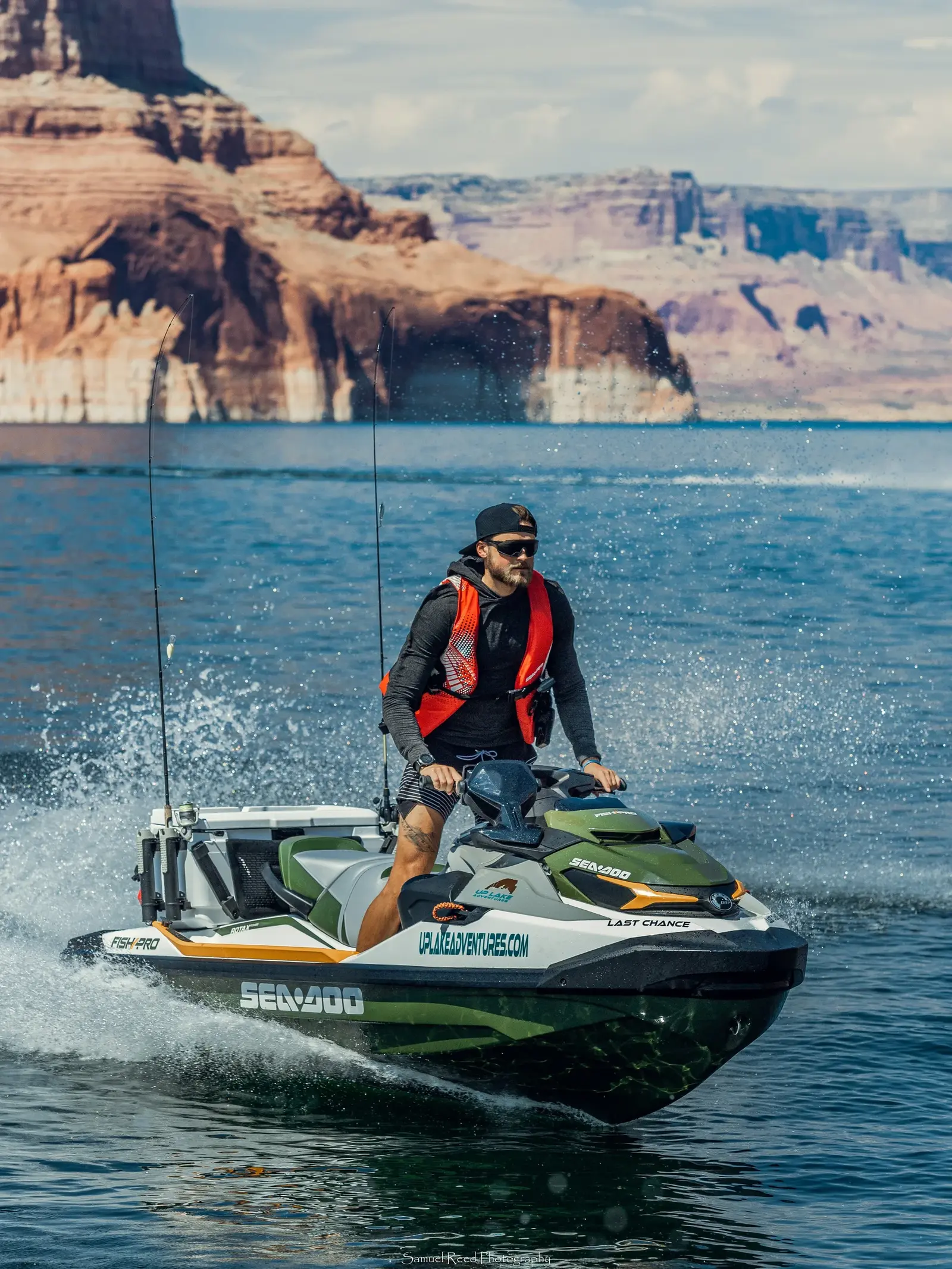 A man wearing a black cap, sunglasses, and a black and red life vest is riding a green and white Sea-Doo jet ski on a body of water with rock formations in the background.