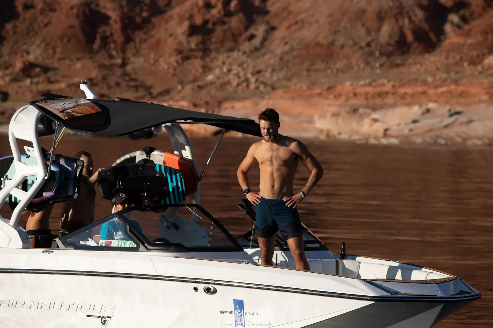 A shirtless man standing on a white speedboat with an orange and black life jacket, two other men are seated inside, and a rocky landscape with water in the background.