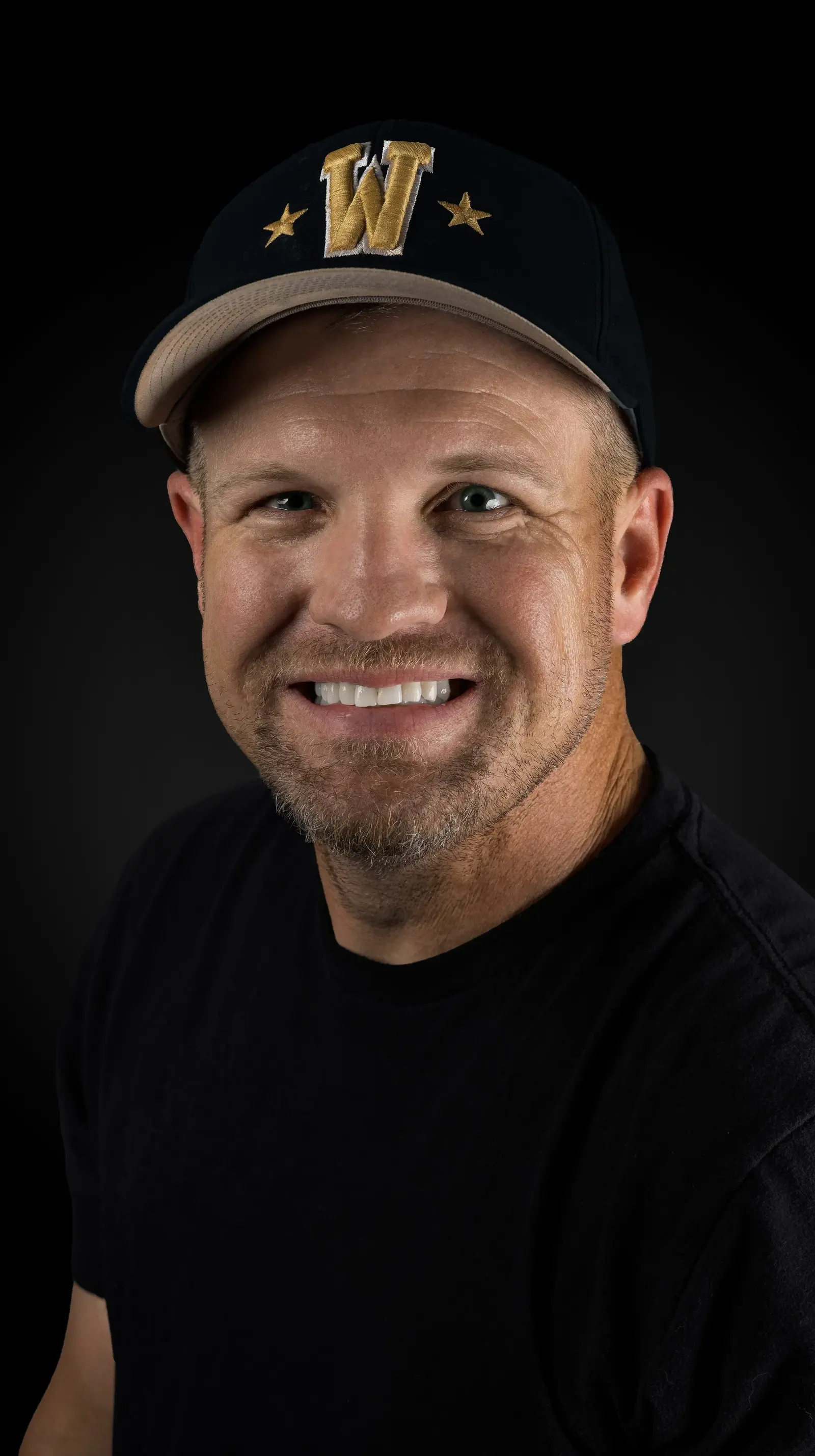 Sam Reed smiling in a black shirt and black baseball cap with a gold W, against a dark background.